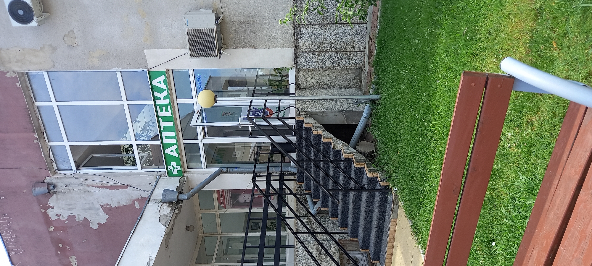 This image shows the entrance to a pharmacy in Glavinitsa, Bulgaria. The pharmacy has a green sign with white text that says "АПТЕКА," which translates to "pharmacy." The entrance is accessed by a set of outdoor stairs with black metal railings. To the left of the stairs, there is a poster displayed in the window of an adjacent building, featuring a young girl and text related to "lavamed." An air conditioning unit is mounted on the exterior wall to the right of the pharmacy entrance. A street lamp with a yellow globe stands near the entrance. The area is surrounded by a grassy lawn, and a portion of a brown wooden bench is visible in the foreground. The building's facade appears weathered, with some peeling paint.