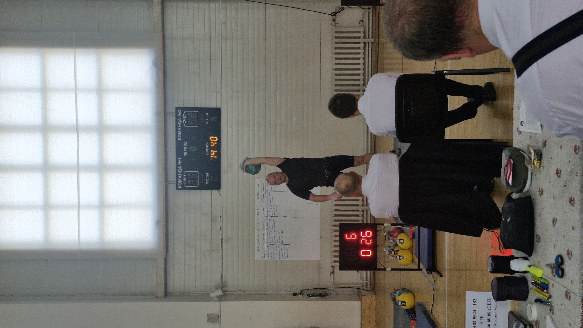 The image shows the interior of a gymnasium or large hall, possibly in Лобаново, Russia. A man dressed in black is performing a side plank exercise while holding a green medicine ball overhead. Three people are seated on chairs, observing him. A scoreboard on the wall displays the time as "14.40". Another digital display near the floor reads "0:26". There is a window on the left side of the frame, and various objects are on a table in the foreground. A printed sheet with writing in Russian is attached to the wall.