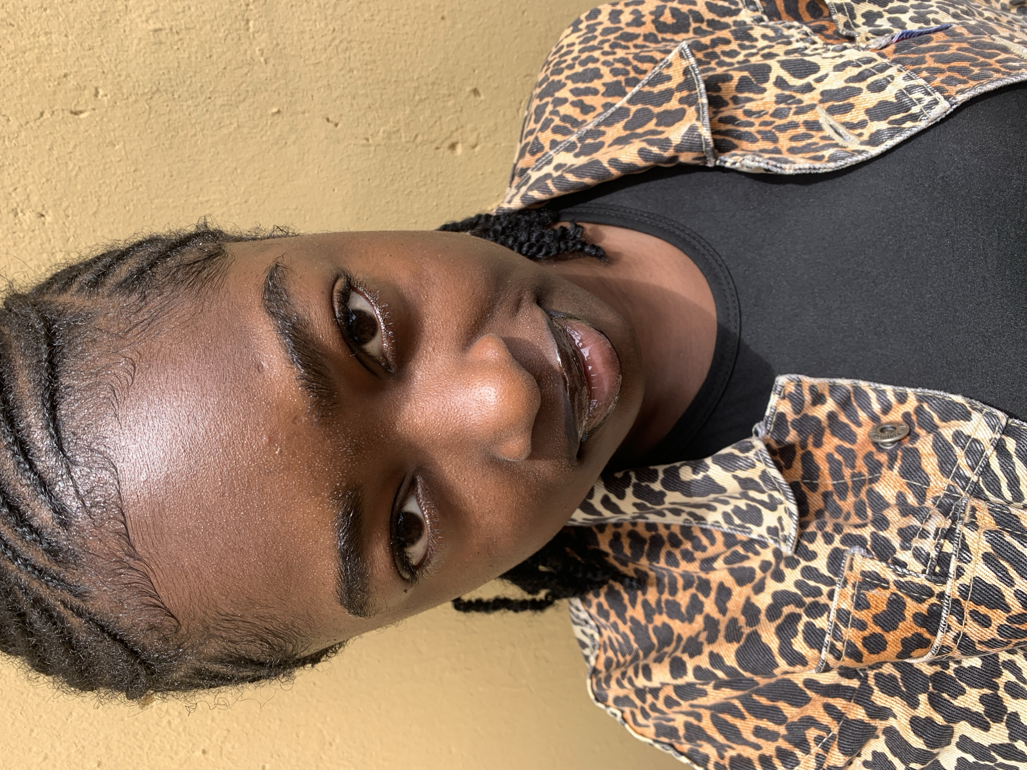 A close-up, slightly high-angle portrait features an individual with dark skin, facing directly towards the camera. The person has cornrow braids styled neatly with intricate baby hairs along the hairline, exhibiting a subtle sheen. Their dark brown eyes maintain direct eye contact with the viewer. Eyebrows are dark and well-defined. The nose has a broad base. Lips are full and appear to be coated with a clear or dark gloss, with the mouth slightly open. The individual's complexion shows natural highlights, particularly on the forehead and nose. They are wearing a black crew-neck top underneath a denim-style jacket with a distinctive leopard print pattern in shades of black, orange-brown, and beige. Visible details of the jacket include stitching, a collar, and a metal button on a chest pocket flap. The background consists of a uniformly textured wall painted in a mustard-yellow hue, consistent with a stucco or concrete surface. The scene is illuminated by natural light, casting subtle shadows, suggesting an outdoor setting in Rafin Par, Nigeria.