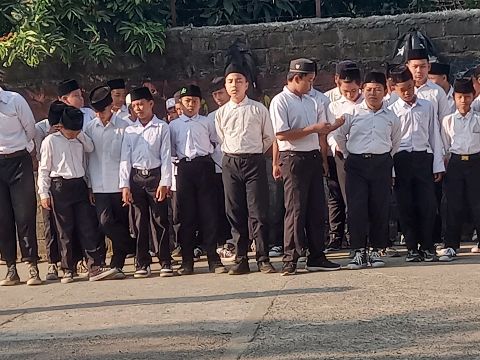 In the city of Perampuan, Indonesia, a group of young males, appearing to be students, are lined up outdoors. They are dressed in white long-sleeved shirts and black pants, with many wearing black skullcaps or kufis. The students are positioned in rows, facing forward, suggesting a formal gathering or assembly. The environment includes a textured concrete ground, a dark brick wall in the background, and lush green foliage overhead. The lighting suggests it is daytime, possibly morning or afternoon, with sunlight casting shadows on the ground. The students' expressions and postures are varied; some appear attentive, while others have their heads down or are looking elsewhere. The scene captures a moment of organized activity within a school or community setting.