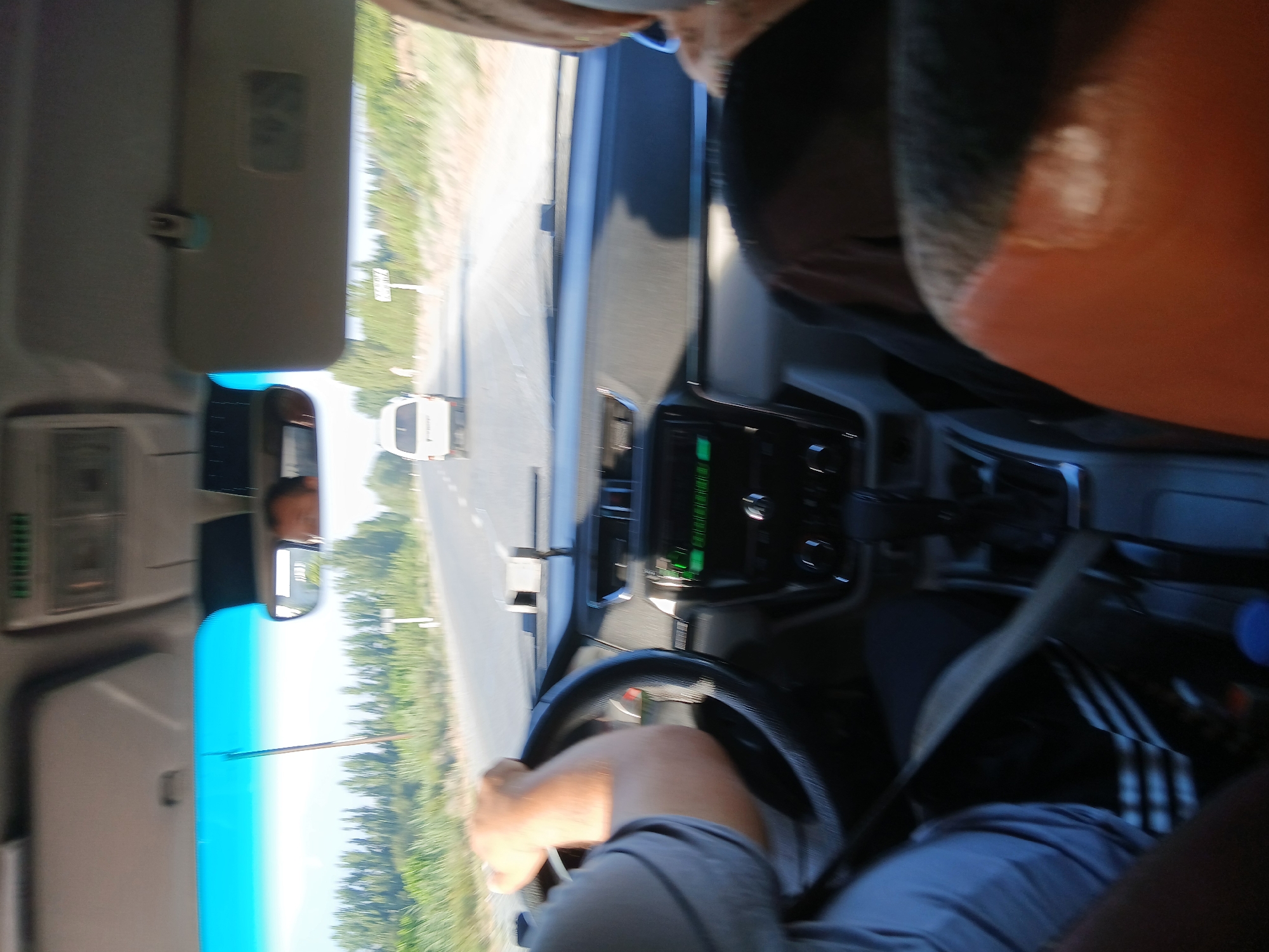 The image shows the interior of a car driving on a paved road. The driver's hand is visible on the steering wheel, and the dashboard with a radio is in the foreground. A white van is visible ahead on the road. The surrounding environment appears to be rural, with trees and dry vegetation lining the road. The image is blurry, indicating motion. Based on the visual cues, the scene is consistent with a road in the vicinity of Khankiz, Uzbekistan, a region known for its arid landscapes and rural roads.