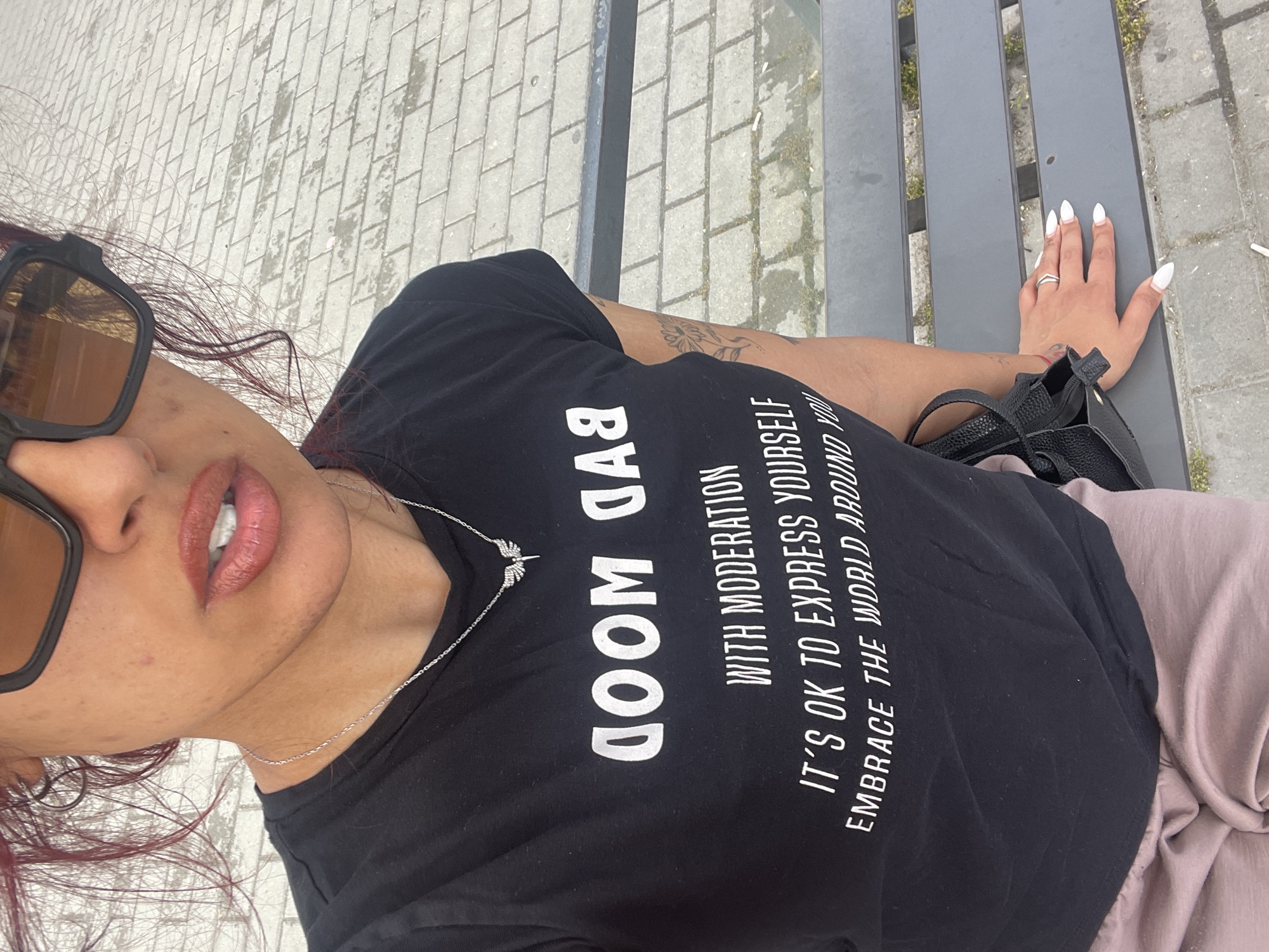 An individual is seated outdoors on a dark gray metal slatted bench in an Unknown city, Unknown. The person, visible from the upper chest to mid-thigh, wears a black short-sleeved t-shirt featuring white text. The topmost line of text is mirrored, reading "DOOM DAB" (intended as "BAD MOOD"), followed by "WITH MODERATION", "IT'S OK TO EXPRESS YOURSELF", and partially "EMBRACE THE WORLD AROUND YOU". A thin silver chain necklace with a small, sparkling angel wing pendant is worn. Large, black-framed sunglasses with dark lenses obscure the upper facial features. The lips are parted, revealing some upper teeth, and are coated with a glossy, reddish-brown color. Strands of reddish-brown hair are visible around the face. A small reddish mark is present on the right cheek. A light brown or pinkish pair of loose-fitting pants covers the lower body. The person's left hand (viewer's right) rests flat on the bench, displaying long, pointed white fingernails and a silver ring on the ring finger. A faint floral tattoo is visible on the right forearm (viewer's left), adjacent to a black handbag resting on the lap or beside the body. The ground consists of light gray rectangular pavers with sparse green growth between them.