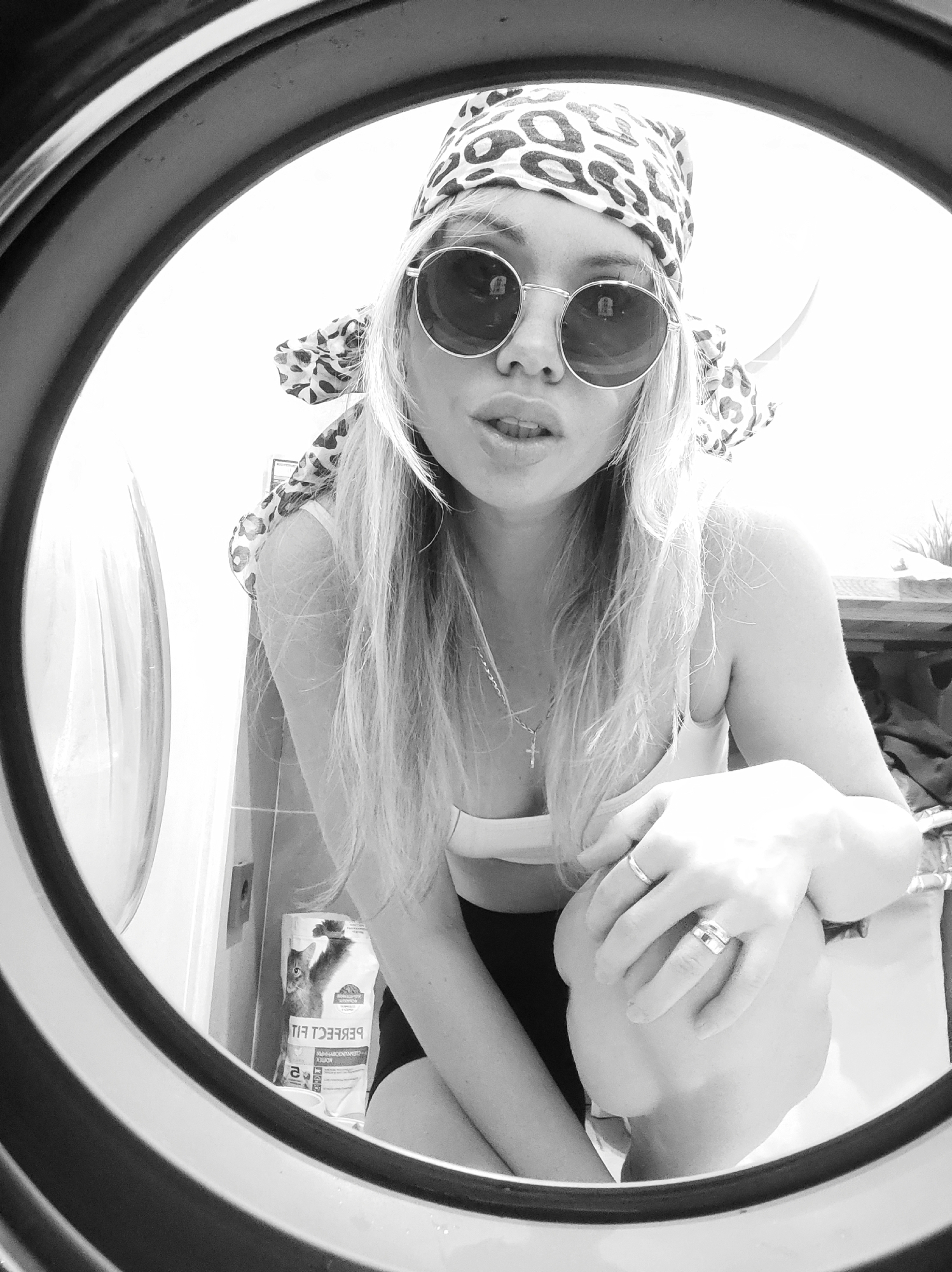 The image is a black and white selfie taken from inside a washing machine or dryer. A young woman with long, light hair and wearing a patterned headscarf and round sunglasses is looking into the lens. She is wearing a light-colored top and dark shorts. On the wall behind her, a package of cat food is visible. The exact location in Votkinsk, Russia, cannot be determined from the image.
