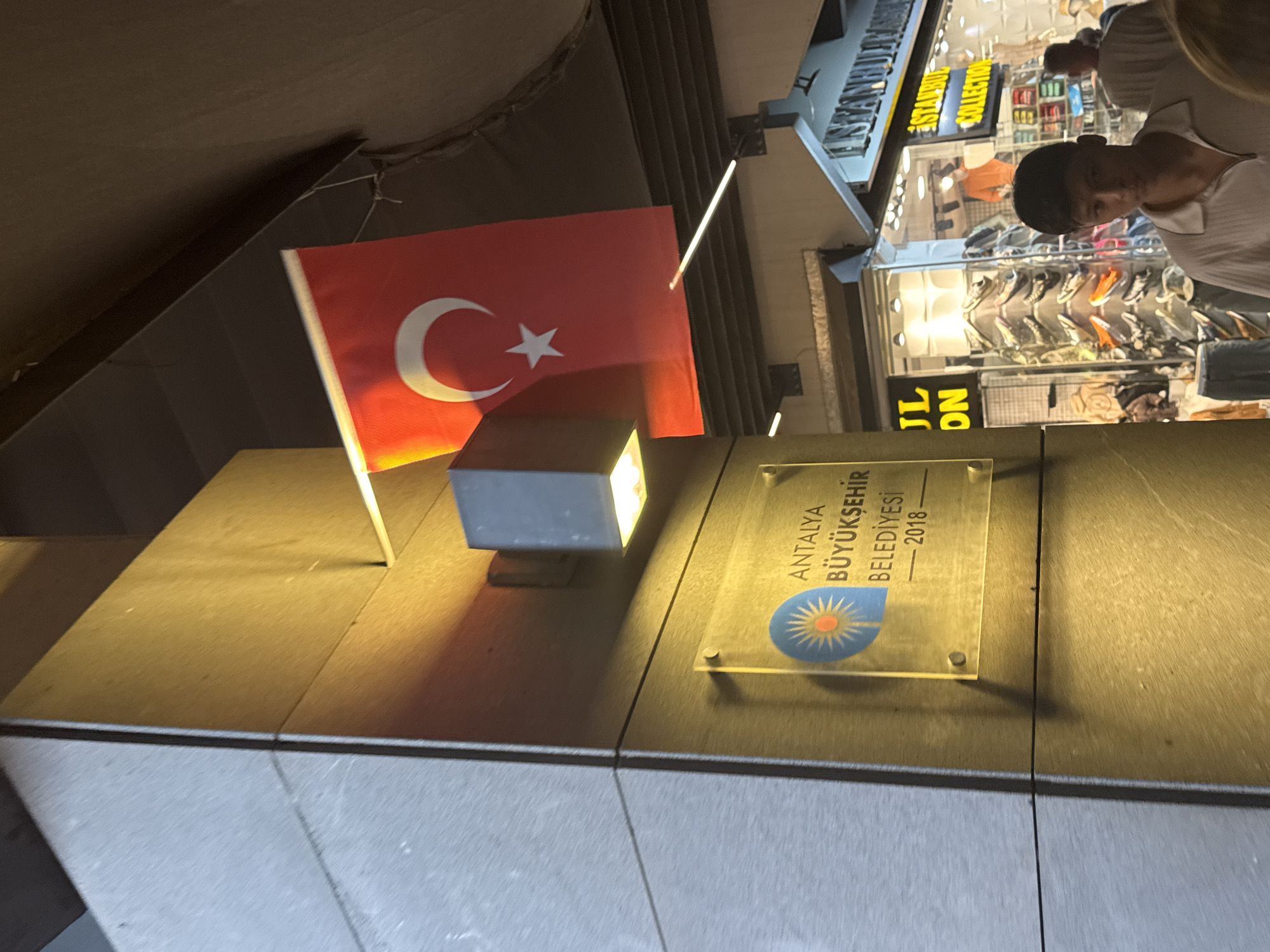 The image depicts a street scene in Antalya, Turkey, with a focus on a plaque and a Turkish flag. The plaque, made of clear acrylic, is mounted on a dark, textured wall and features the text "ANTALYA BÜYÜKŞEHİR BELEDİYESİ - 2018" along with a circular emblem of a sun. To the right of the plaque, a Turkish flag with a white star and crescent on a red background is displayed.
In the background, several shopfronts are visible, indicating a commercial area. One shop has a sign that reads "JL ON", and another displays "ISTANBUL COLLECTION". The shops appear to be selling clothing and accessories, with mannequins and merchandise visible through the windows. A young man in a white polo shirt is standing in the foreground, looking towards the camera. Other people are blurred in the background. The lighting suggests it is either dusk or evening, with artificial lights illuminating the shops and the plaque.