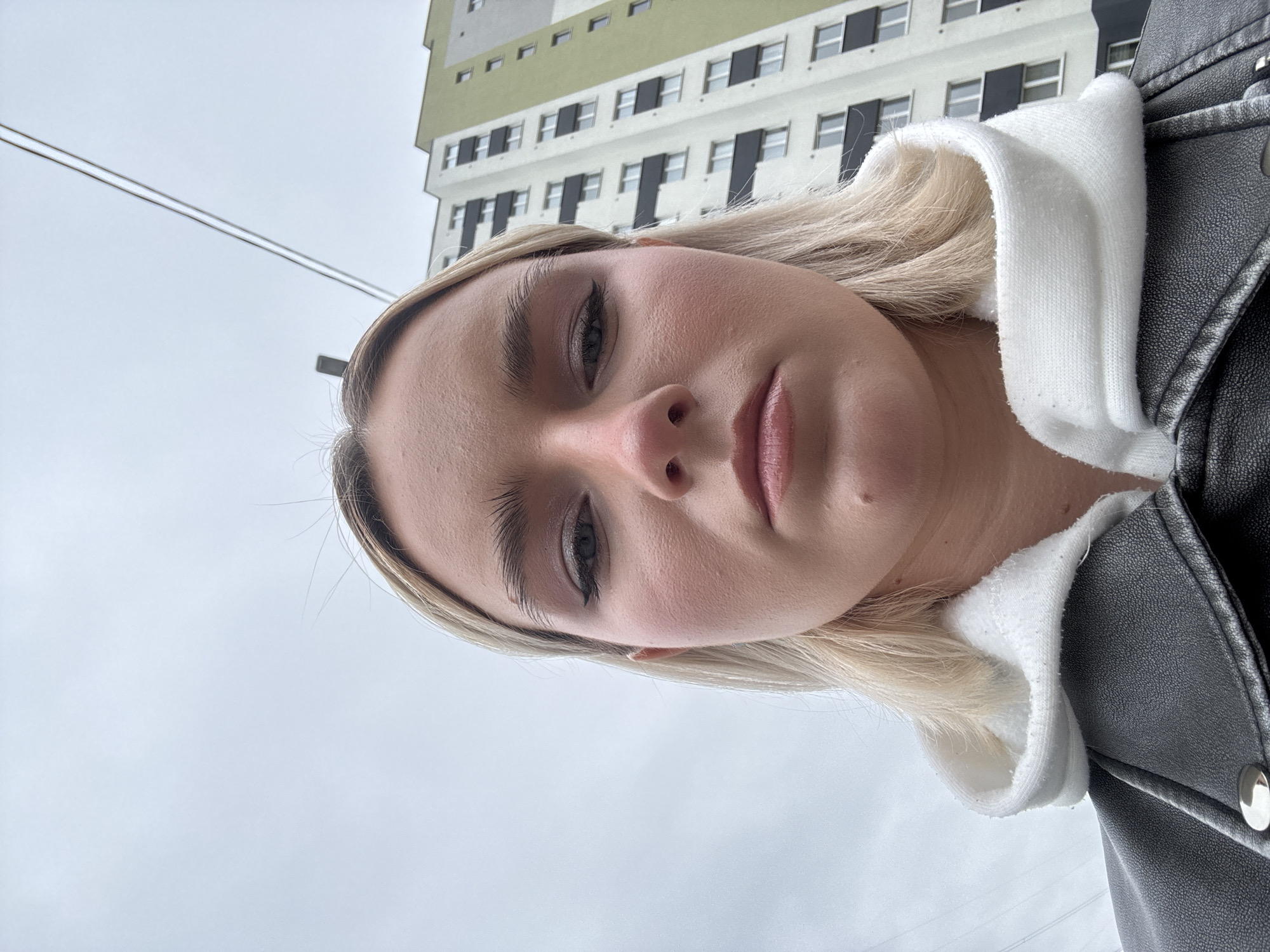 A close-up, selfie-style photograph depicts a fair-skinned person with light blonde hair wearing a white hooded sweatshirt and a black leather jacket. The background features a multi-story building with white and green panels and black window frames, indicating an urban environment. Above the person, there are visible power lines. The location is indicated as Unknown, Russia.