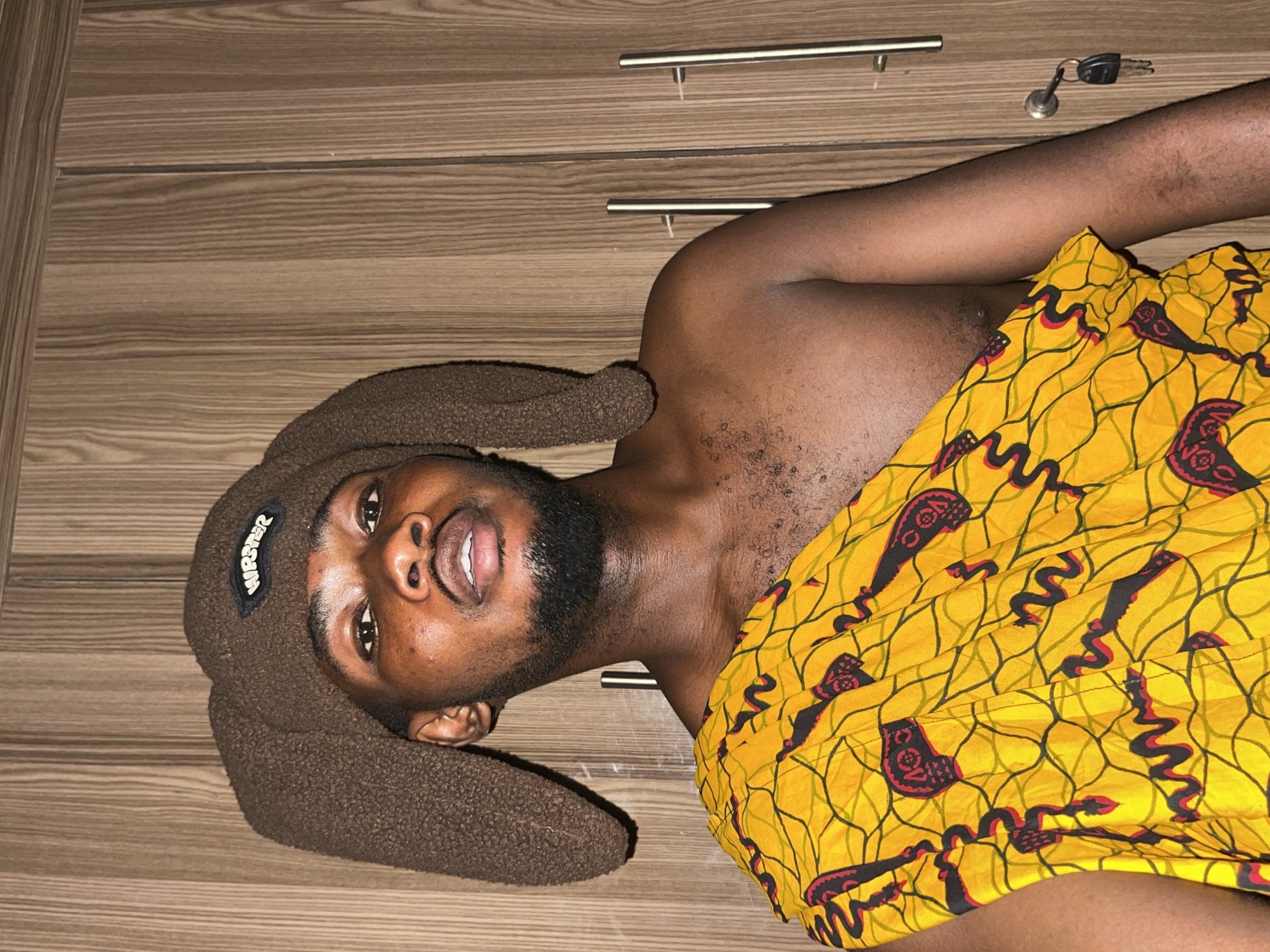 A young man with dark skin and a beard is captured lying on his back, looking directly into the camera with a relaxed, slightly open-mouthed expression. He is wearing a distinctive brown, fuzzy hat with long, floppy ears, reminiscent of a bunny or teddy bear. A small patch on the hat displays the white text "HUSTLER". His upper torso is bare, while his lower body is covered by a vibrant, bright yellow fabric featuring a bold, repeating pattern of black and red abstract shapes, possibly stylized snakes or traditional motifs. This appears to be a type of traditional African print fabric, such as Ankara.
The man is positioned against a light brown wooden surface, which resembles a dresser or wardrobe with a horizontal grain. On this surface, two silver-colored metal drawer handles are visible, along with a set of keys – a silver key and a black fob – resting near his arm. The scene is set indoors, under what appears to be artificial lighting. The overall impression is casual and playful, with the hat and patterned fabric adding a distinctive cultural and personal touch. The location context suggests this image was taken in Unguwar Gabas, Nigeria.