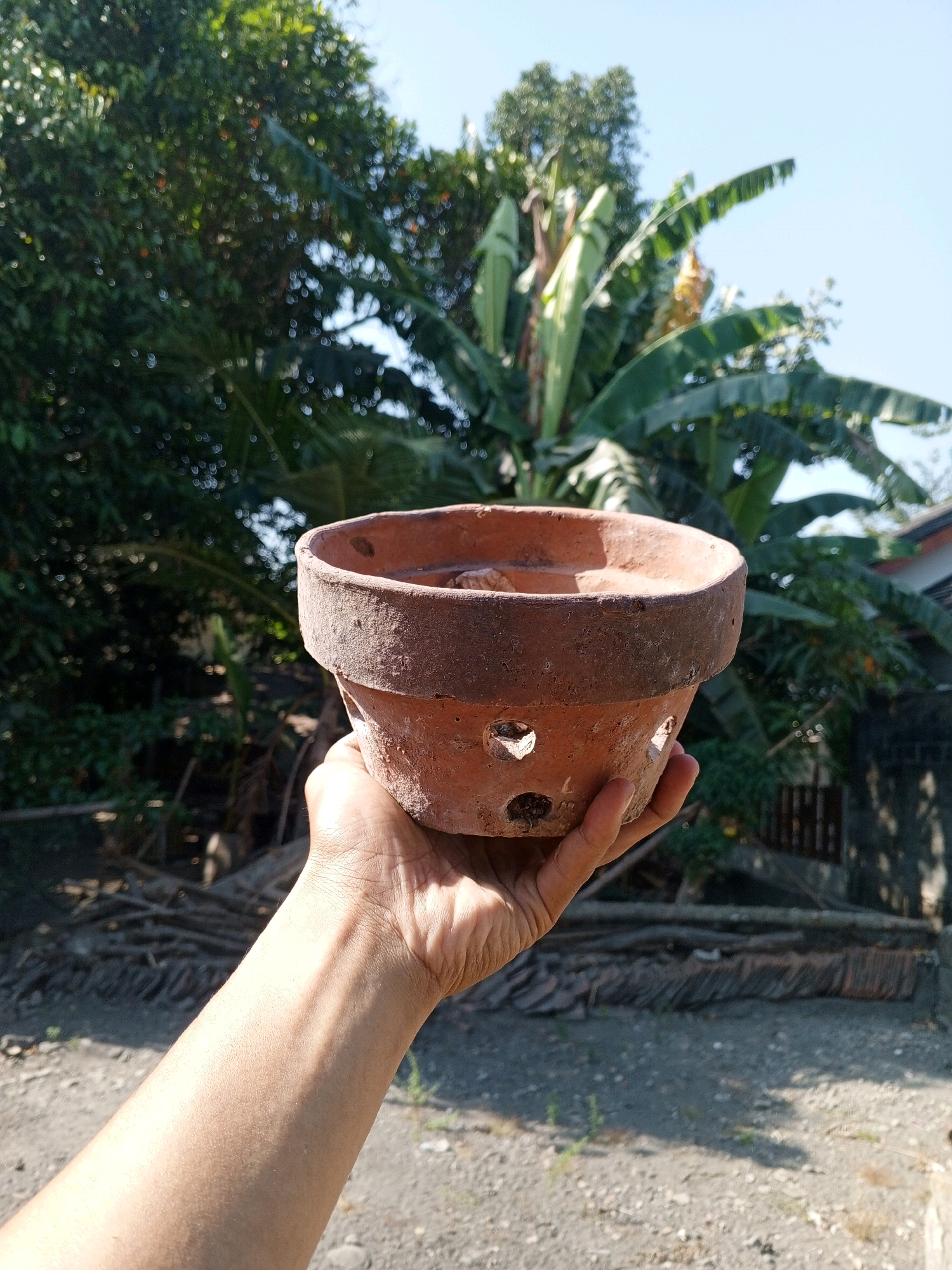 A person's hand is holding a reddish-brown clay pot with ventilation holes. The pot appears to be empty or filled with a small amount of soil. In the background, there are lush green trees, including banana trees, and a clear blue sky. The ground in the foreground is a mix of dirt and gravel. The scene suggests an outdoor setting, likely in a rural or suburban area. The presence of banana trees indicates a tropical or subtropical climate, consistent with Bantul, Indonesia, a regency in Yogyakarta Province.