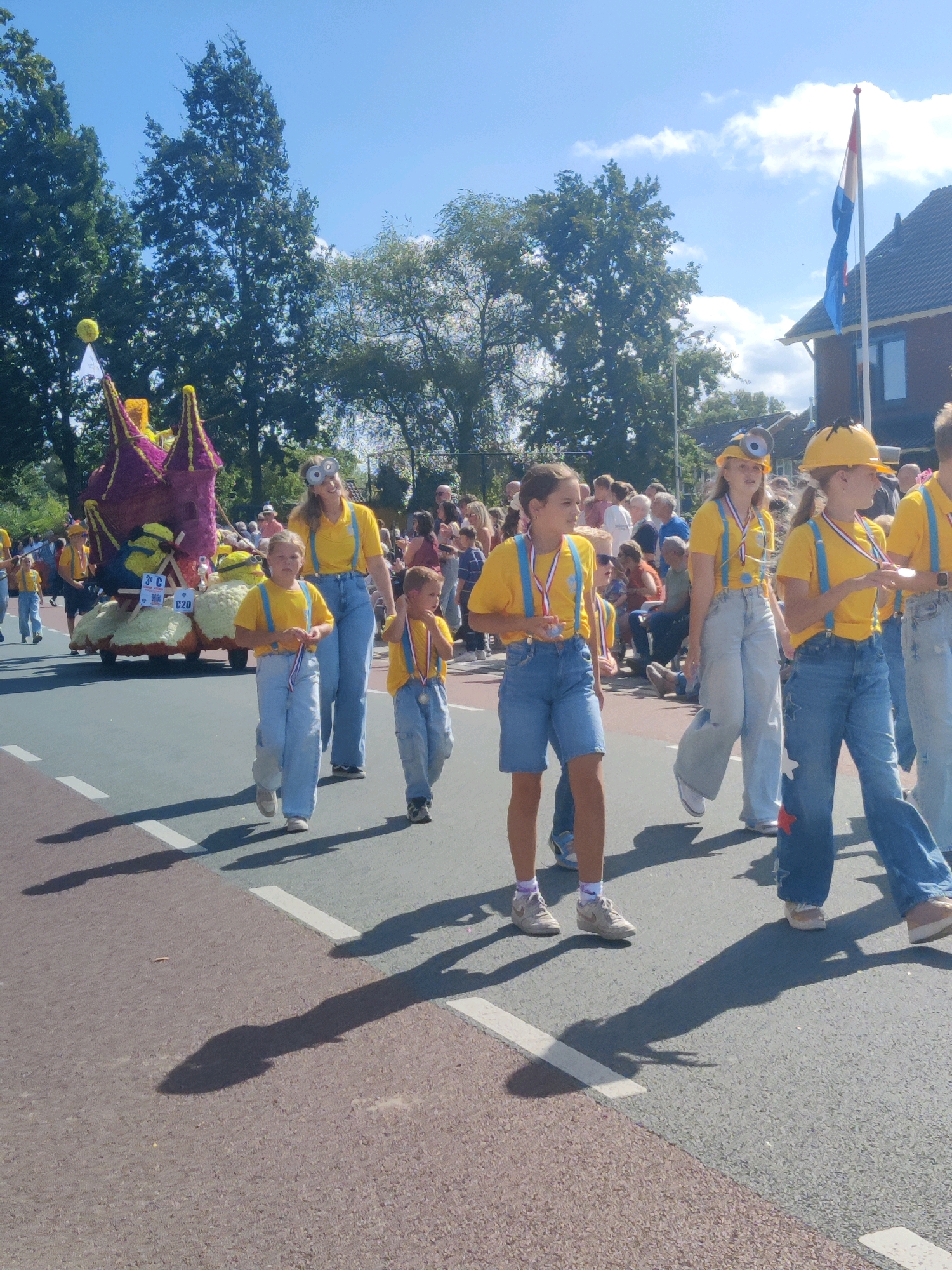 In Winterswijk, Netherlands, a parade is taking place on a sunny day. The foreground features a group of children dressed as Minions, with yellow shirts, blue overalls, and goggles. They are walking in single file on a paved street, some carrying medals. Behind them, a float decorated with yellow and purple floral arrangements resembles a castle. Spectators are visible on the sidewalk and in the background. A Dutch flag flies on a flagpole near a building. The overall atmosphere suggests a festive community event.