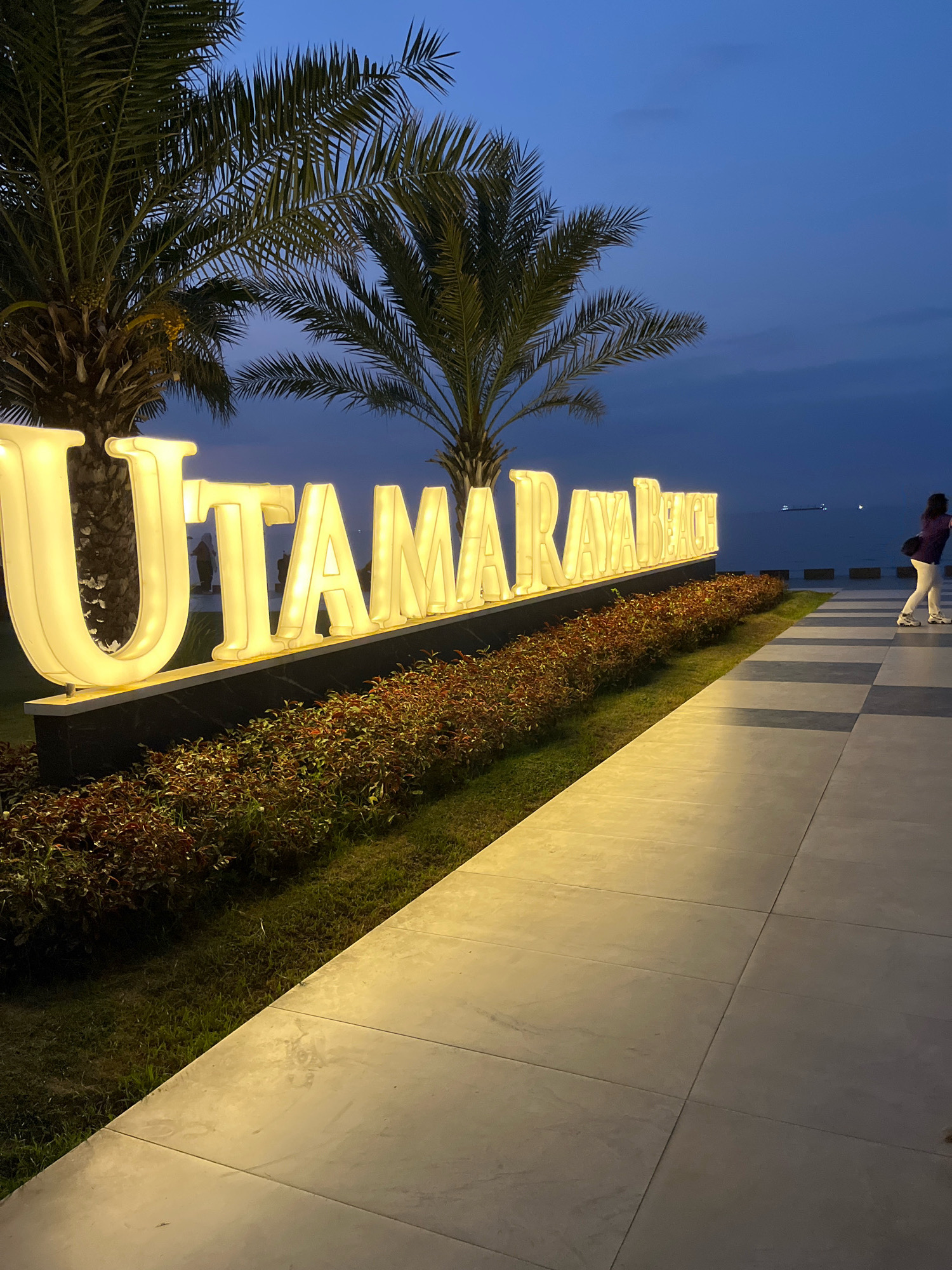 In Banyuglugur, Indonesia, at dusk, a large, illuminated sign reads "UTAMARAYA BEACH". The sign is lit with warm yellow lights and is flanked by palm trees. A paved walkway, with a checkered pattern of light and dark gray tiles, runs along the right side of the sign. A person is walking away from the viewer on the walkway. In the background, the ocean meets the twilight sky, with a distant ship visible on the horizon. The foreground is a strip of green grass and ornamental plants.