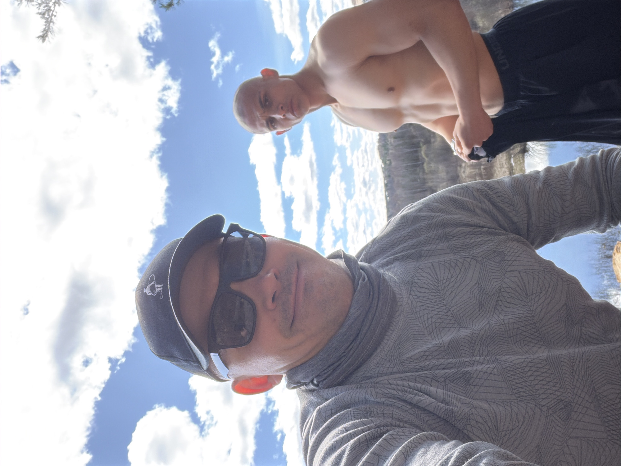 A selfie image taken outdoors on a sunny day with white clouds in the blue sky. The image features two males. The primary subject is wearing a black baseball cap with a white logo, sunglasses, and a patterned gray long-sleeved shirt. He is positioned in the foreground and facing the camera. Behind him is a second male, shirtless and wearing black athletic shorts labeled "UNDER ARMOUR." He is partially obscured and his pose appears casual.
The background suggests a natural environment, possibly near a body of water, with trees faintly visible in the distance. There is no concrete evidence to link the image to Izhevsk, Russia.