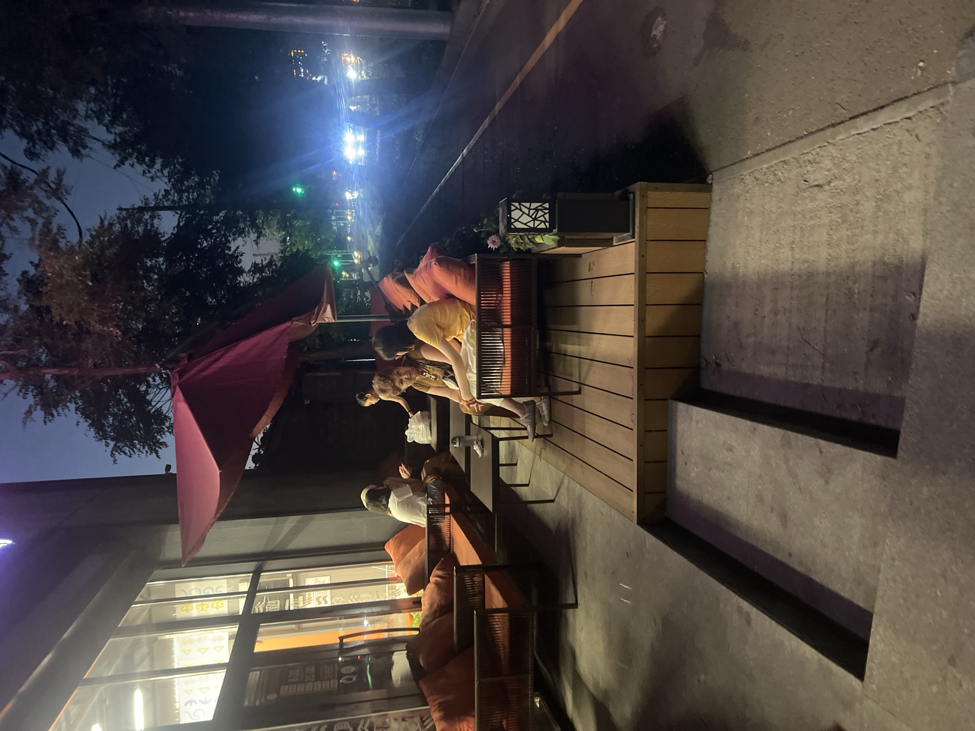 The image depicts an outdoor seating area of a restaurant or cafe in Bishkek City, Kyrgyzstan, at dusk. Several people are seated at tables and on a low platform furnished with orange cushions. In the foreground, concrete steps lead down from the seating area to a sidewalk. A wooden deck forms the seating platform, and a planter with a decorative light fixture sits on its edge. Beyond the seating area, a road is visible with car headlights and traffic lights illuminated in the twilight. Trees line the roadside, and the building facade of the establishment features large windows with interior lighting. One group of people appears to be engaged in conversation, while another individual is seated alone. The overall scene suggests a casual dining or social gathering setting.