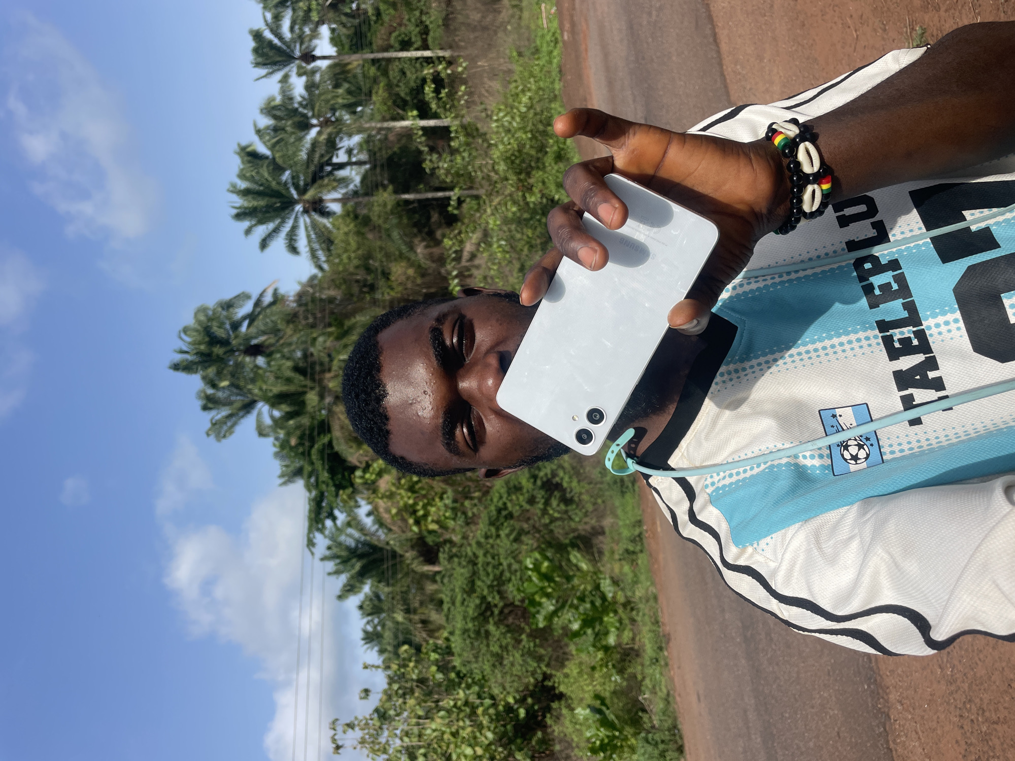 A man stands on an asphalt road in Ogbe, Nigeria, holding a light-colored smartphone to his ear. He is wearing a white and light blue jersey with black trim, adorned with a soccer emblem and the word "TALEPLU." A bracelet made of black beads, cowrie shells, and beads with red, yellow, and green stripes is visible on his wrist. The background features lush green vegetation with tall palm trees under a partly cloudy sky.