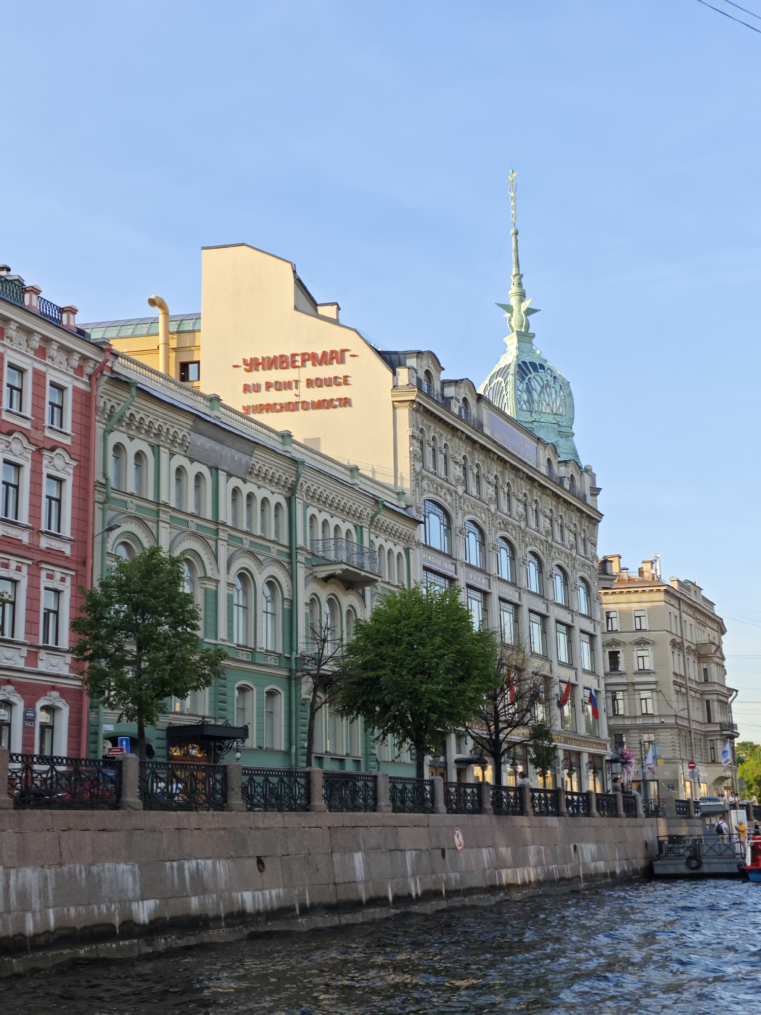 The image shows a view of the Singer House building in Saint Petersburg, Russia, as seen from the Griboyedov Canal. The Singer House, with its distinctive green dome and spire, is the central architectural feature. In front of it are several other historic buildings with ornate facades, painted in colors such as pink, green, and beige. A stone embankment with a decorative black metal railing lines the canal. Several trees are visible along the embankment. The water in the canal is dark and rippled. A small boat is docked at the embankment on the right side of the image. The sky is clear and blue. The overall scene depicts a picturesque urban landscape along a canal.