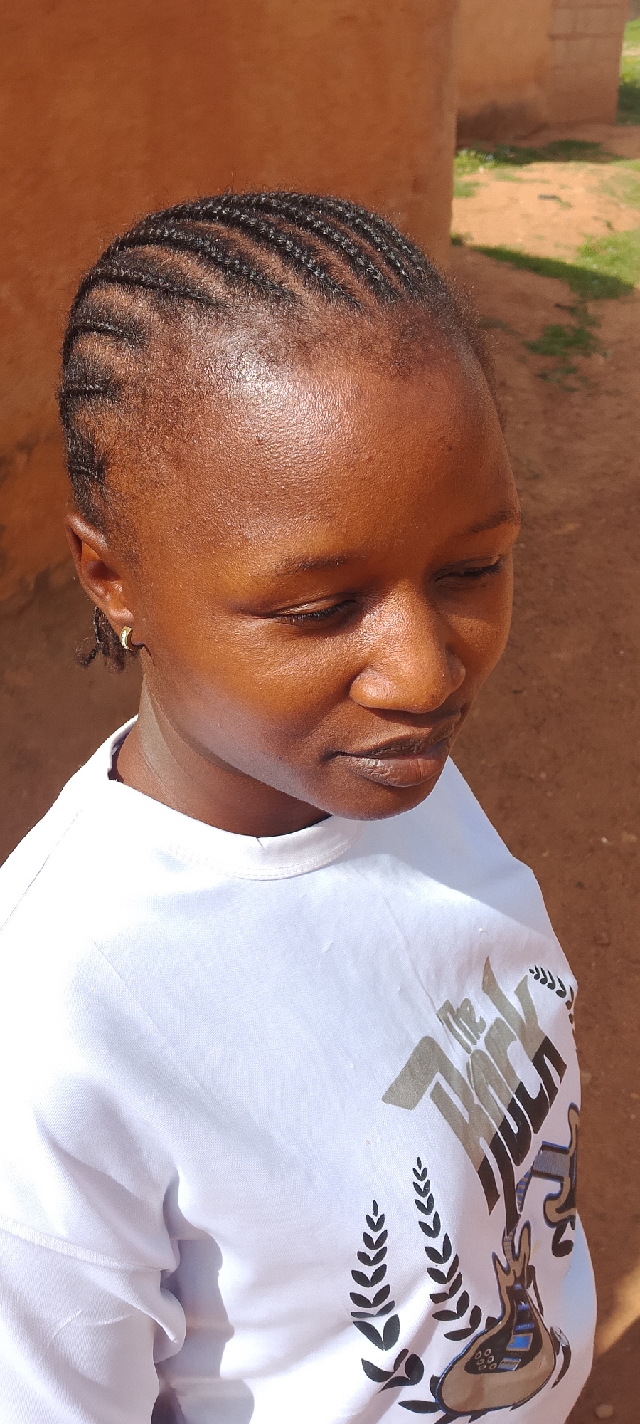 A woman with cornrow braids and wearing a white t-shirt with a graphic design stands outdoors. The location appears to be in Jos, Nigeria, based on the background featuring what may be a mud-brick building and the individual's attire and features. The woman is looking down with a pensive expression. She has small gold hoop earrings. The area behind her appears to be a dirt or reddish soil terrain. The lighting suggests it's a sunny day. The graphic on the t-shirt includes the words "THE ROCK" along with laurel or wheat stalk designs and a stylized guitar.