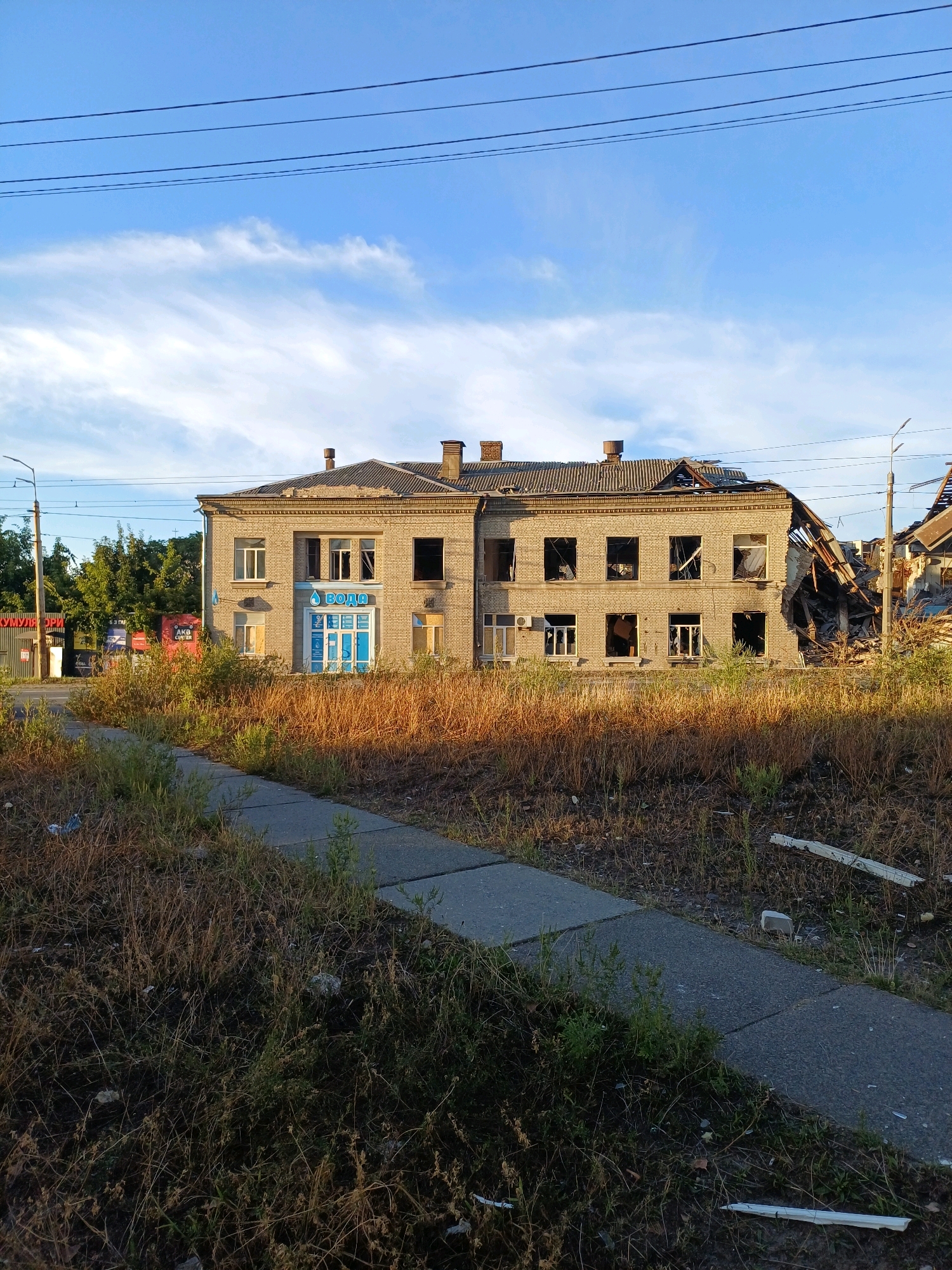 This image depicts a two-story brick building with significant damage, located in Kyiv, Ukraine. The right side of the building shows extensive destruction, with a collapsed roof and walls exposing the interior structure and debris. The windows on both floors are largely broken or missing. The building appears to be an abandoned or damaged commercial establishment, with a sign above the entrance of the left side of the building that reads "ВОДА" (water) in blue lettering. The surrounding environment is overgrown with dry grass and weeds, and a concrete sidewalk runs in the foreground. The sky above is blue with scattered wispy clouds. Power lines are visible across the top of the frame. The overall scene suggests a post-conflict or disaster environment.