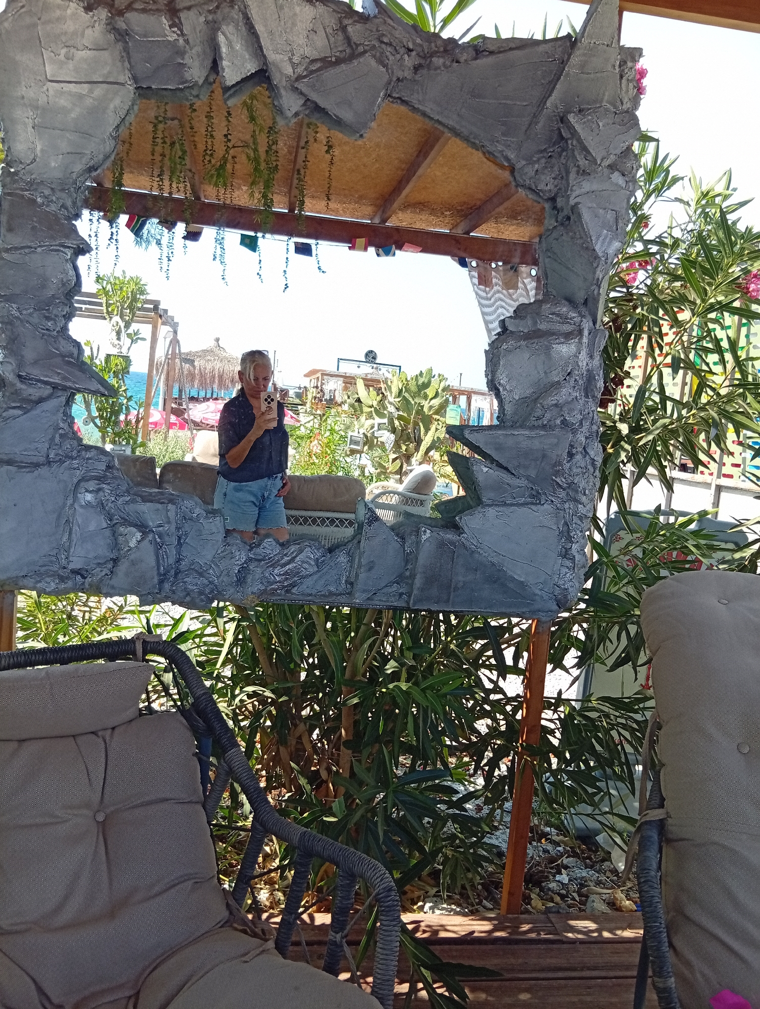 The image is a selfie taken by a woman standing outdoors in what appears to be a resort or restaurant setting, likely in Trikomo, Northern Cyprus. The woman is facing a mirror, which is framed by a rough, grey, rock-like structure. She is wearing a dark blue short-sleeved shirt and light blue denim shorts, holding a smartphone up to take the picture. Her hair is light-colored and pulled back.
Behind her, reflected in the mirror, is a view of the sea, a beach with sun loungers and umbrellas, and some buildings. The background of the actual scene shows lush green foliage, including plants with pink flowers, and parts of outdoor furniture, specifically a cushioned chair in the foreground. The overall ambiance suggests a sunny day in a coastal location. The unique mirror frame suggests a decorative element of the establishment.