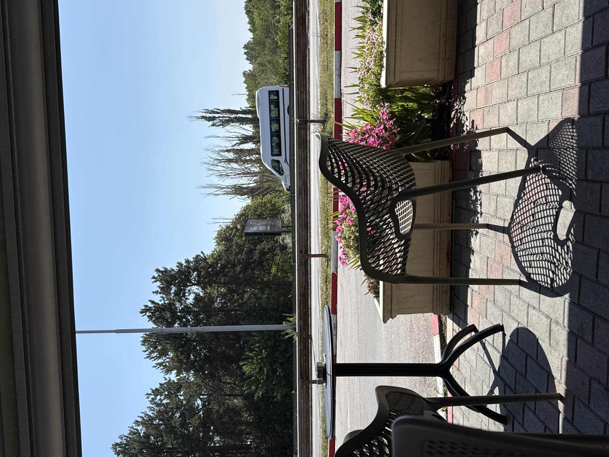 This image depicts an outdoor seating area, likely a cafe or restaurant patio, overlooking a road with a white van in motion. In the foreground, there are two dark-colored, modern plastic chairs, one prominently in focus with a complex shadow cast on the brick-patterned ground. A black round table with a metal object, possibly an ashtray, is also visible. Adjacent to the chairs are two rectangular planters filled with vibrant pink flowers and greenery.
The background reveals a paved road with a red and white striped curb, bordered by a metal guardrail. Beyond the road, a dense line of green trees stands against a clear blue sky. A tall utility pole is present on the left side of the frame, and a white van with blue trim is driving on the road in the middle ground. The overall scene is well-lit, suggesting it was taken during daylight hours under sunny conditions. The location is identified as Unknown, Unknown.