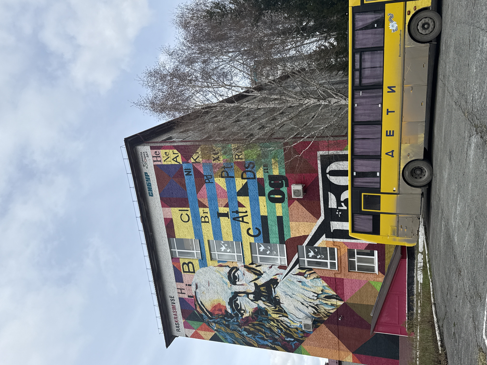 The image shows the side of a colorful building in Tobolsk, Russia, featuring a large mural. The mural includes a portrait of Dmitri Mendeleev, the creator of the periodic table, as well as various chemical elements. Text on the mural reads "СИБУР ХОРОШИХ ДЕЛ" (SIBUR, good deeds), "ФОРМУЛА ЧЕЛОВЕКА" (formula of man), and "RASKRASIMVSË" (let's paint everything). A yellow bus is parked adjacent to the building, with the Cyrillic word "ДЕТИ" (children) written on its side. Trees are visible in the background, and the sky is partially cloudy. The setting appears to be an urban street or parking area.