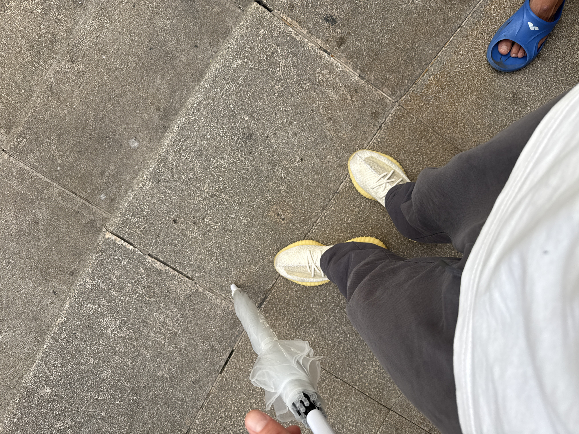 This image captures a first-person perspective of someone standing on a set of stone stairs in Haizhu District, China. The person is wearing dark gray pants, a white t-shirt, and light-colored athletic shoes. To the left, the person is holding a clear plastic umbrella. Further down the stairs, a blue sandal is visible on another person's foot. The stone steps appear worn and slightly damp.