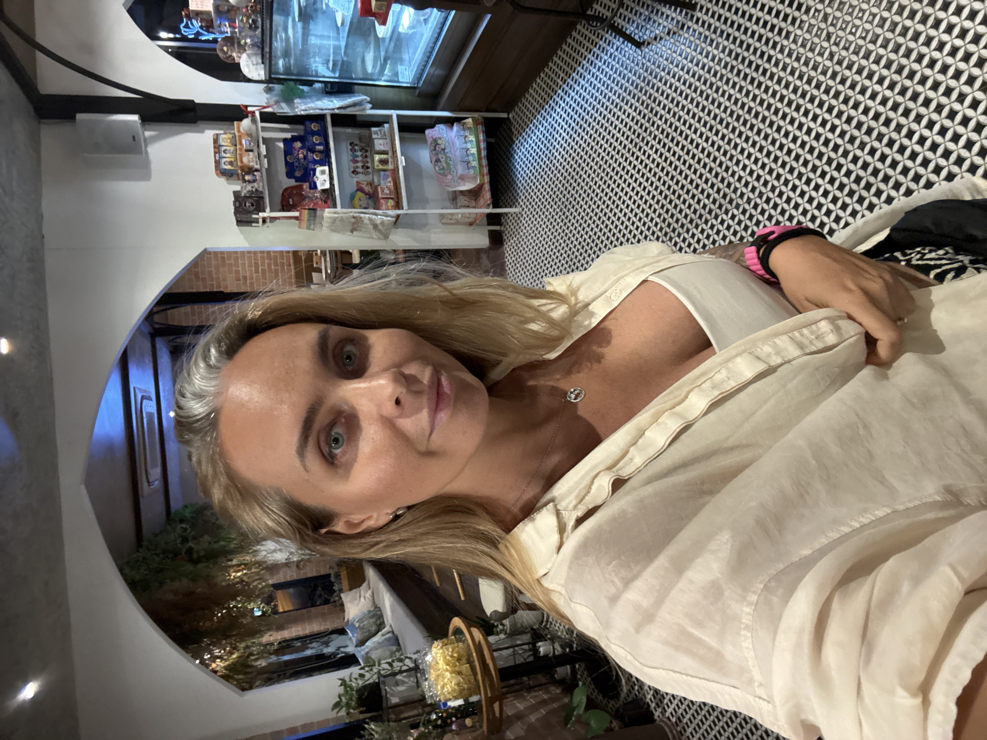 A woman with blonde hair and blue eyes is taking a selfie indoors. She is wearing a cream-colored shirt and a white tank top, with a necklace and a pink and black bracelet on her wrist. She is standing in front of a black and white patterned floor. Behind her, there is a display shelf with various items, and a glass display case. To her left, there is a cushioned bench with pillows and plants. The location is a cafe in the city of Ko Samui District, Thailand.