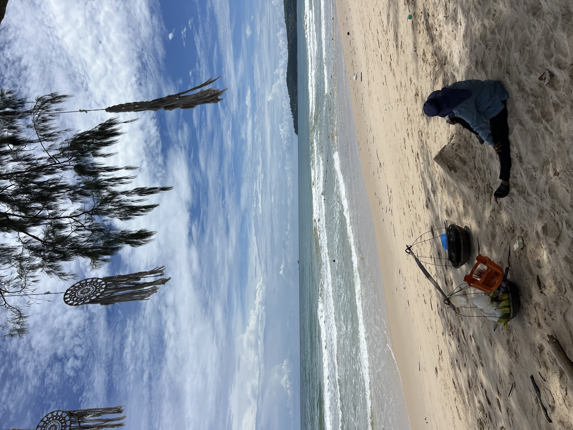A person is sitting on a sandy beach, looking out at the ocean. The person is wearing a blue shirt, black pants, and a blue head covering. In front of the person, there is a pole with baskets of fruit, and further out, a shopping basket. The beach is lined with trees, and the ocean has small waves. The sky is blue with scattered clouds, and there are dreamcatchers hanging from the trees.