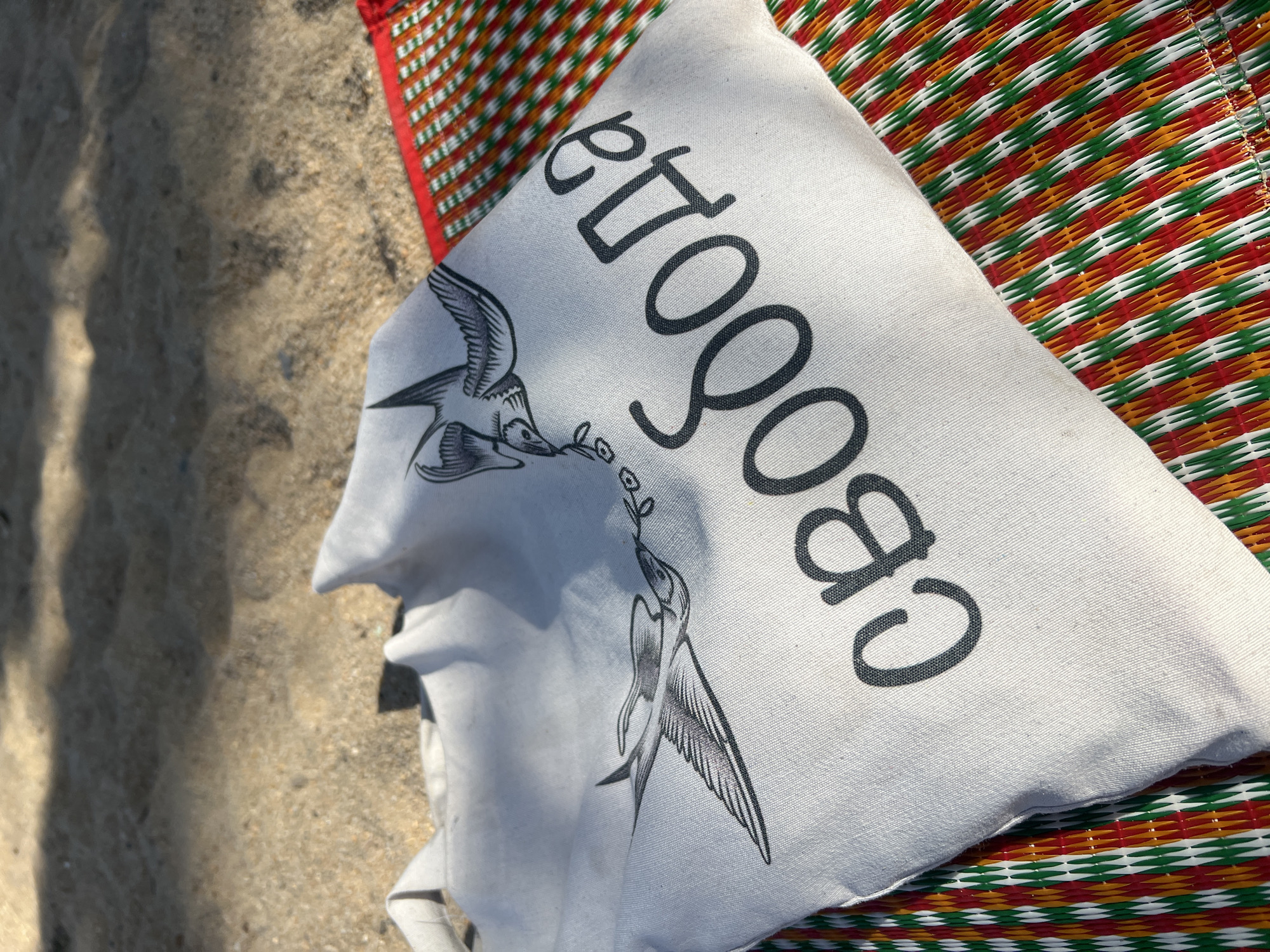 The image depicts a partially visible sandy beach and a woven mat with stripes of red, orange, green, and white. Resting on the mat is a white cloth bag with two drawn birds holding a small sprig of leaves in their beaks. Printed in black on the bag is the word "свобода," a Russian word that means "freedom". The setting appears to be outdoors in the Nha Trang Ward area of Vietnam, based on the beach environment.