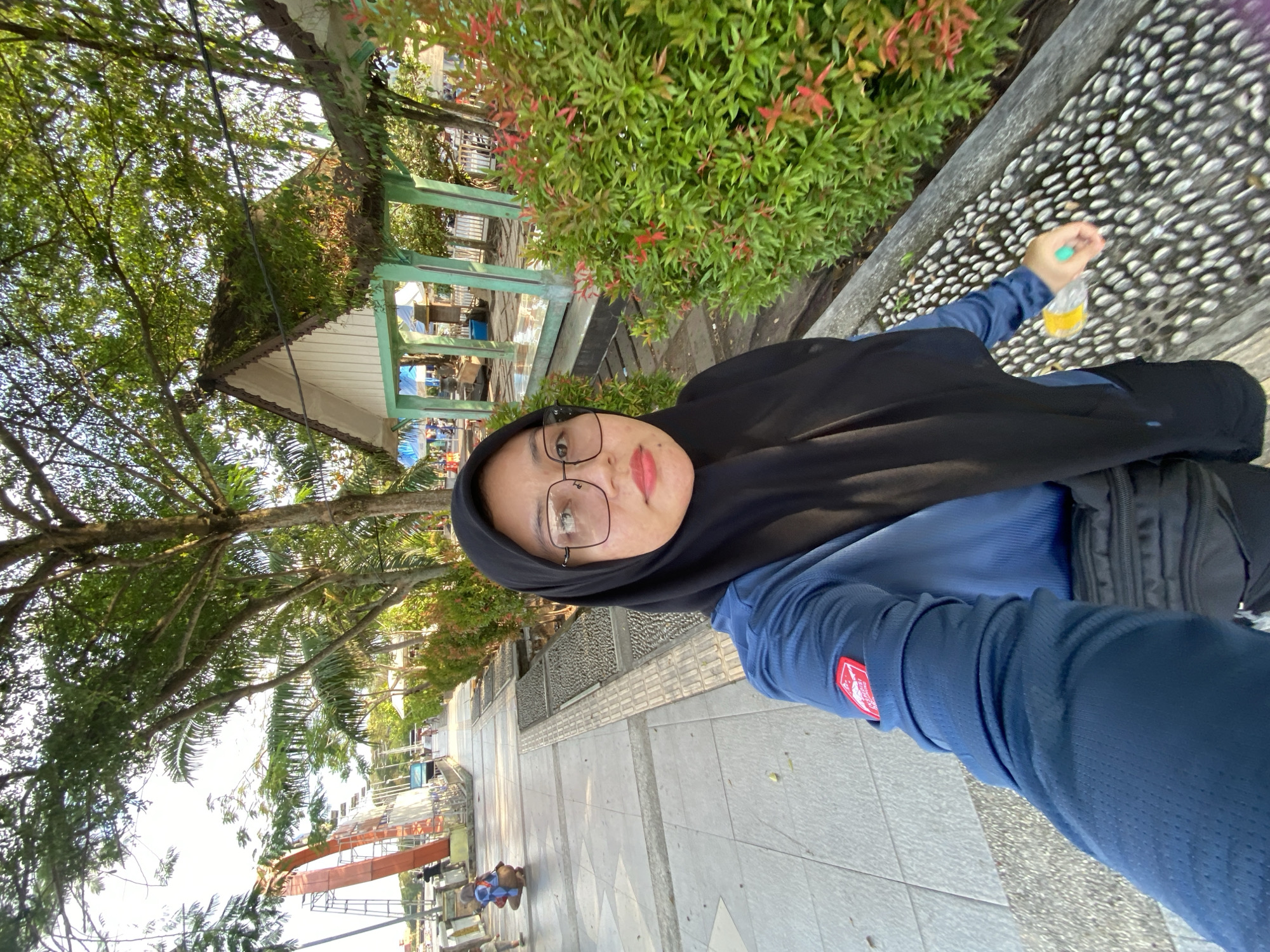 A young woman wearing a blue long-sleeved shirt, black hijab, and glasses is taking a selfie outdoors. She is standing on a sidewalk made of grey tiles and interspersed with textured gray inserts. To her right, there are lush green bushes with red-tipped leaves. In the background, a small pavilion with a green roof and white walls is visible. Further behind, there are trees, a red structure with metal scaffolding, and other people walking along the sidewalk. The scene appears to be located in a park or recreational area within the city of Banjarmasin, Indonesia, possibly near a waterfront given the open space and recreational structures. The lighting suggests it is daytime, with sunlight casting shadows on the ground.