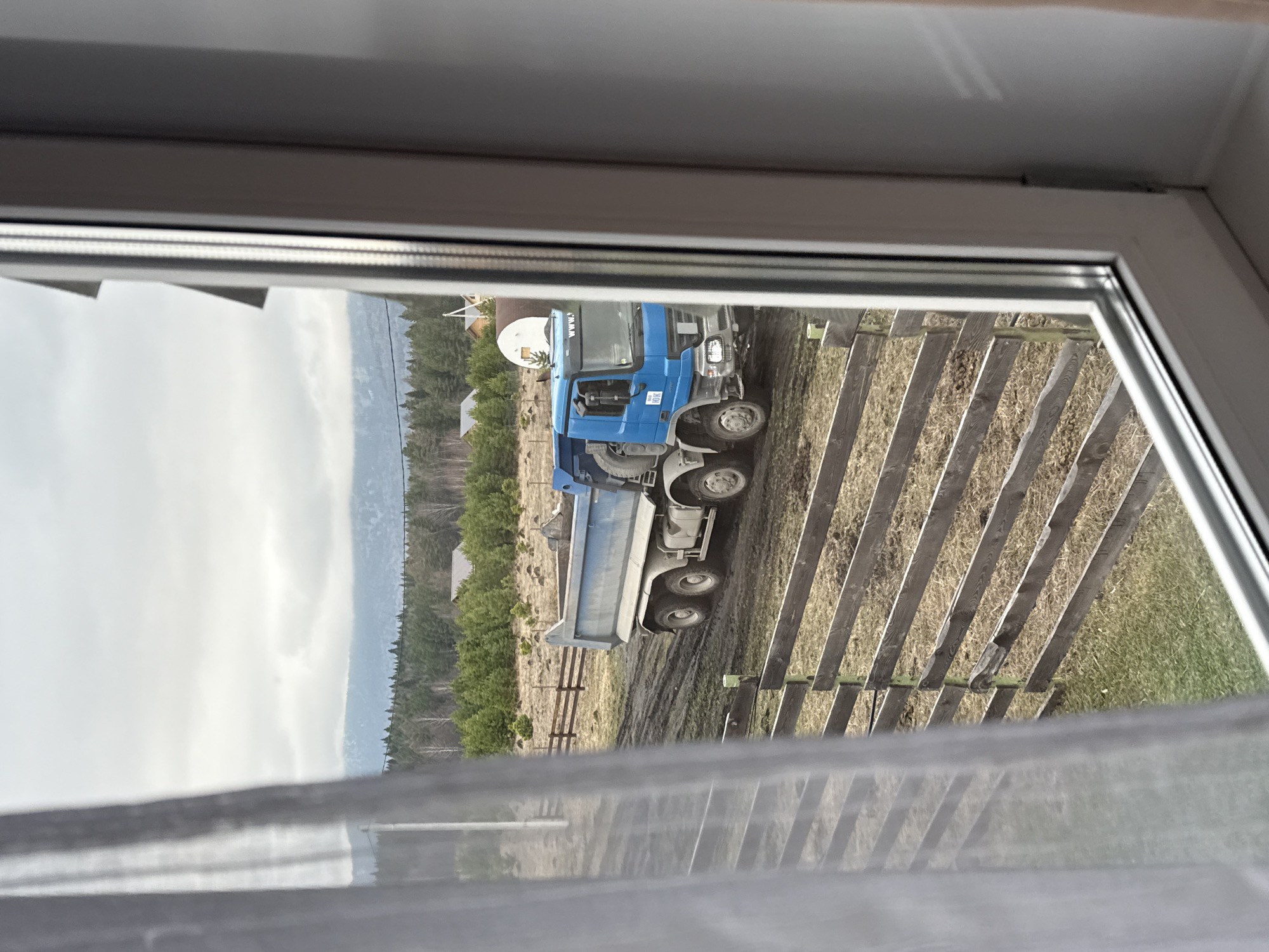 A blue dump truck sits on a muddy lot next to a wooden fence, as viewed through a window. In the background, a forest and mountains are visible. A building with a white satellite dish is also present. The location is Tyulyuk, Russia.