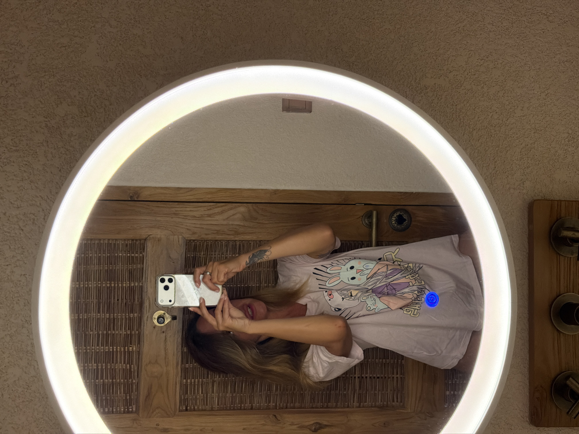 A woman with long, blonde hair is taking a selfie in a circular mirror with a bright LED light. She is wearing a light pink t-shirt with a cartoon design on it. The mirror reflects a wooden door with woven detailing and a handle. The background appears to be a textured wall, possibly in a bathroom. The lighting suggests it is evening or indoors. The image was captured in a location within Mengwi, Indonesia.