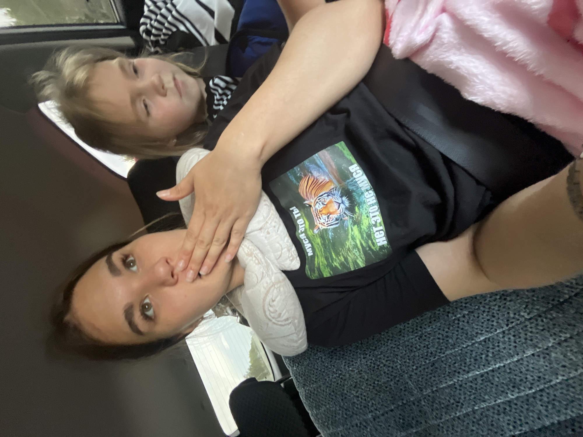 The image shows a woman in the back seat of a car, with a child in the seat behind her. The woman has a snake around her neck and is covering her mouth with her hand. She is wearing a black t-shirt with a graphic of a tiger and text. The child is looking away from the camera. The car appears to be moving, as suggested by the blur of the scenery outside the window. The location is specified as Goldyrevskii, Russia.