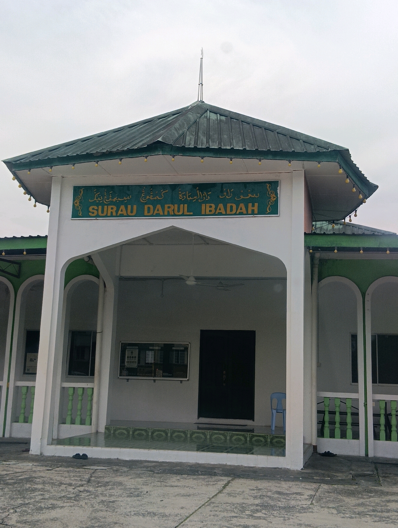 The image shows the front entrance of a building, likely a surau or small mosque, in Kuching, Malaysia. The building has a green, sloping roof with decorative yellow lights strung along its eaves. A white facade features an arched entryway with the words "SURAU DARUL IBADAH" in yellow lettering above the entrance. Arabic script is also visible above the English text. Inside the entryway, a dark double door is visible, and a light blue plastic chair sits to the right of the doorway. Green railing details are present on the sides of the entrance. A bulletin board is mounted on the wall to the left of the entrance. The ground in front of the building is a concrete pavement. A pair of black sandals are placed on the ground near the left side of the entrance. The sky is overcast.