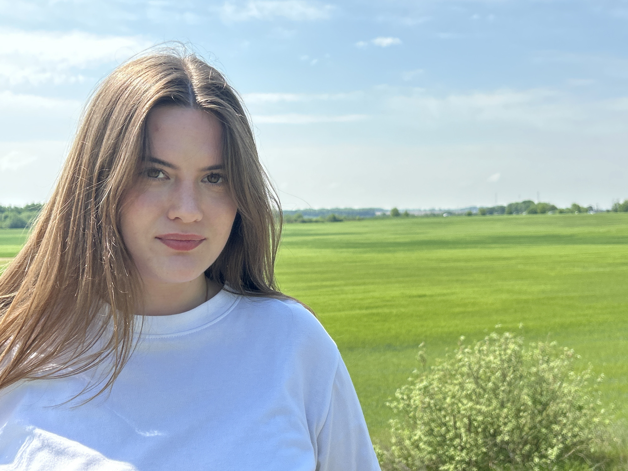 A portrait of a young woman with long brown hair. She is wearing a white t-shirt. The background consists of a green field and blue sky with scattered clouds. The location is Unknown, Unknown.