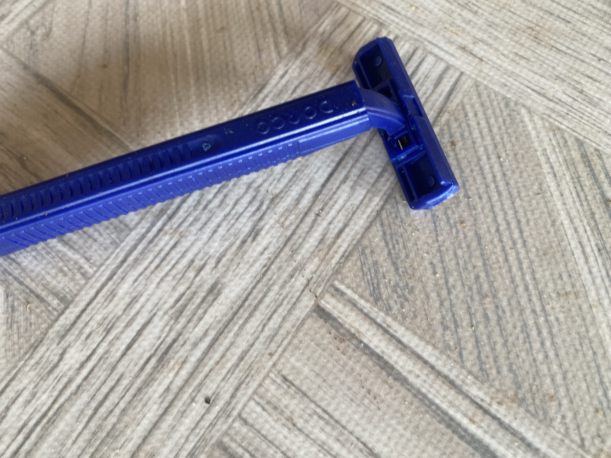 The image shows a blue disposable razor resting on a light-colored tiled surface. The tiles are arranged in a herringbone pattern and have a textured, wood-like appearance. The razor is positioned diagonally, with the handle extending upwards towards the top right of the frame and the head towards the bottom left. The handle has a textured grip and some branding, which appears to be embossed text, is visible. The head of the razor is a bright blue plastic housing for the blades. The surface appears slightly dusty or dirty. The scene is indoors, given the tiled surface, but no further context about the location within Jauro Yinu, Nigeria is provided.