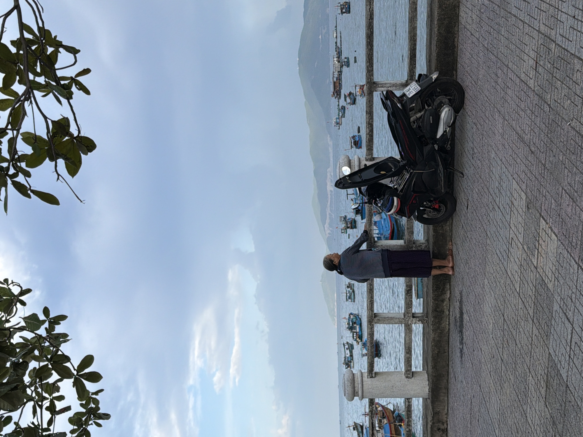 A person stands on a paved promenade, looking out at a body of water filled with numerous small fishing boats. The person is wearing a striped shirt and dark pants. A black scooter is parked on the promenade to the right of the person, with its seat open. The water is calm, and in the distance, a hazy mountain range is visible. Overhanging branches of a tree frame the top of the image. The scene is likely the coastal promenade along Tran Phu Street in Nha Trang, Vietnam, given the characteristic presence of fishing boats and the mountainous backdrop.