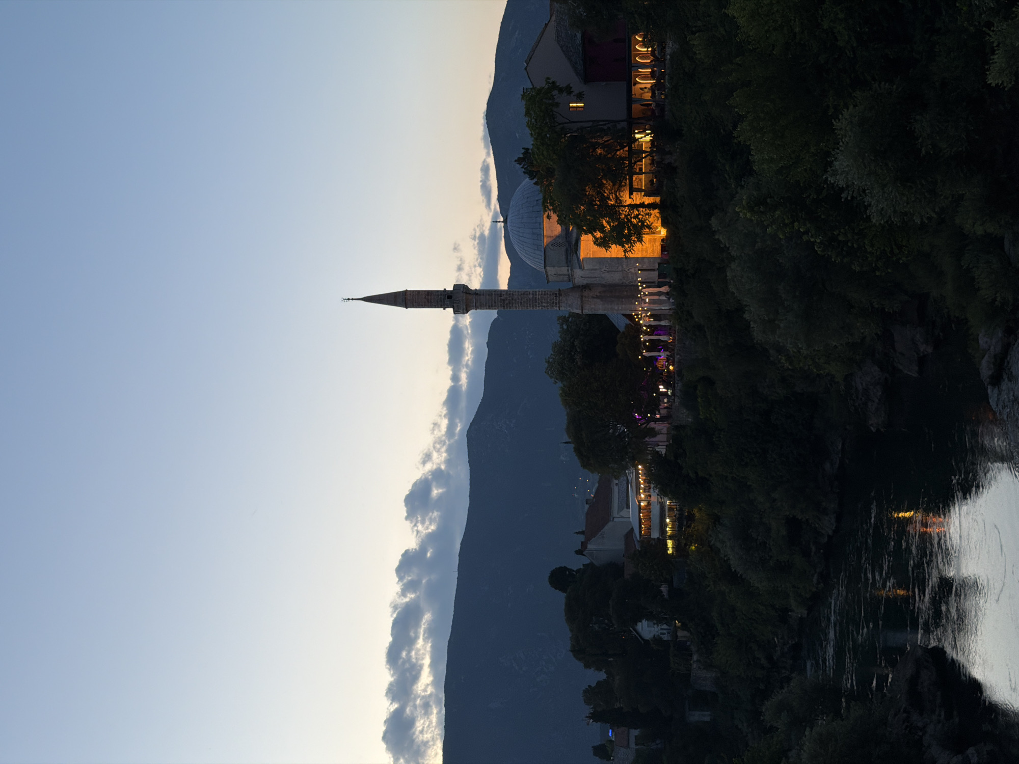 The image depicts a twilight scene in Mostar, Bosnia and Herzegovina, focusing on the Koski Mehmed Pasha Mosque and its surroundings. A tall, slender minaret pierces the sky on the left, adjacent to a dome-shaped structure illuminated by warm, orange light emanating from within the mosque. Below the mosque, a row of illuminated restaurants and cafes line the bank of the Neretva River, their lights reflecting on the water's surface. Dense foliage and trees dominate the foreground and mid-ground, with the dark silhouette of mountains forming the backdrop. The sky is a gradient of soft blues and grays, with scattered clouds near the horizon. No people are clearly identifiable.