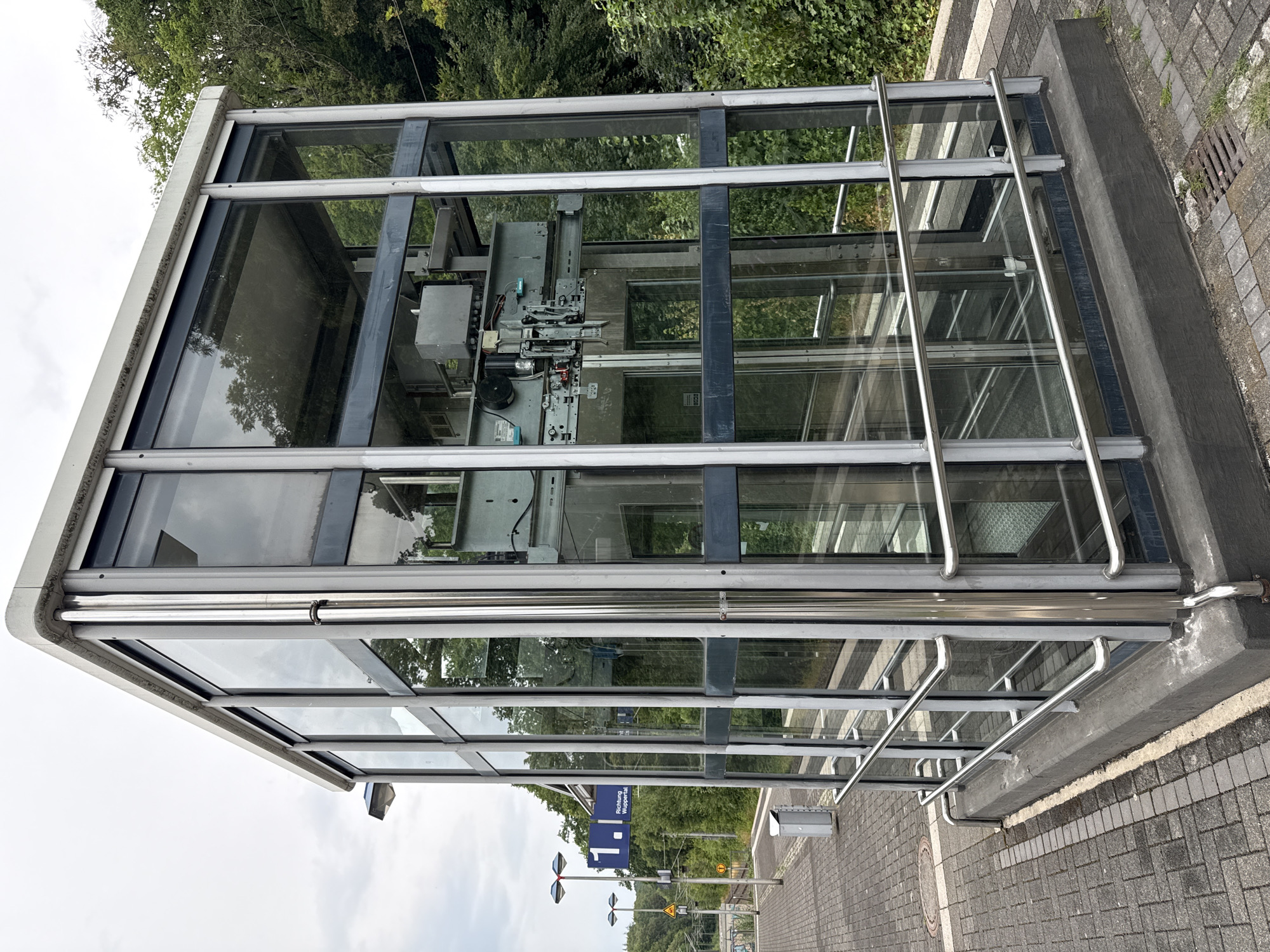 This media file shows an exterior view of a modern glass and metal elevator enclosure, likely at a train station or public transportation hub. The enclosure has a rectangular footprint with a sloped roof, constructed with a metal frame supporting large glass panels. Inside the enclosure, the mechanical components of an elevator are visible, including cables, pulleys, and motor housings. The elevator appears to be a service elevator, judging by the exposed machinery and lack of a passenger car interior visible.
The surrounding environment includes lush green trees and foliage in the background, suggesting a natural or park-like setting. The foreground is a paved area, possibly a sidewalk or platform, made of interlocking grey bricks. A metal railing runs along the exterior of the glass enclosure, providing a safety barrier. A blue and white directional sign with text is visible, reading "Richtung Wuppertal" which translates to "Direction Wuppertal" in German, indicating the location is in Germany and the elevator provides access towards Wuppertal. A small, yellow warning sign with a black symbol is also visible on a pole in the distance.
The lighting suggests it is daytime, with an overcast or cloudy sky visible in the upper portion of the image. The overall impression is one of functionality and modern infrastructure within a potentially scenic or well-maintained public space. There are no people visible in the image. The scene appears to be static, with no specific activity or event taking place other than the presence of the elevator structure itself.