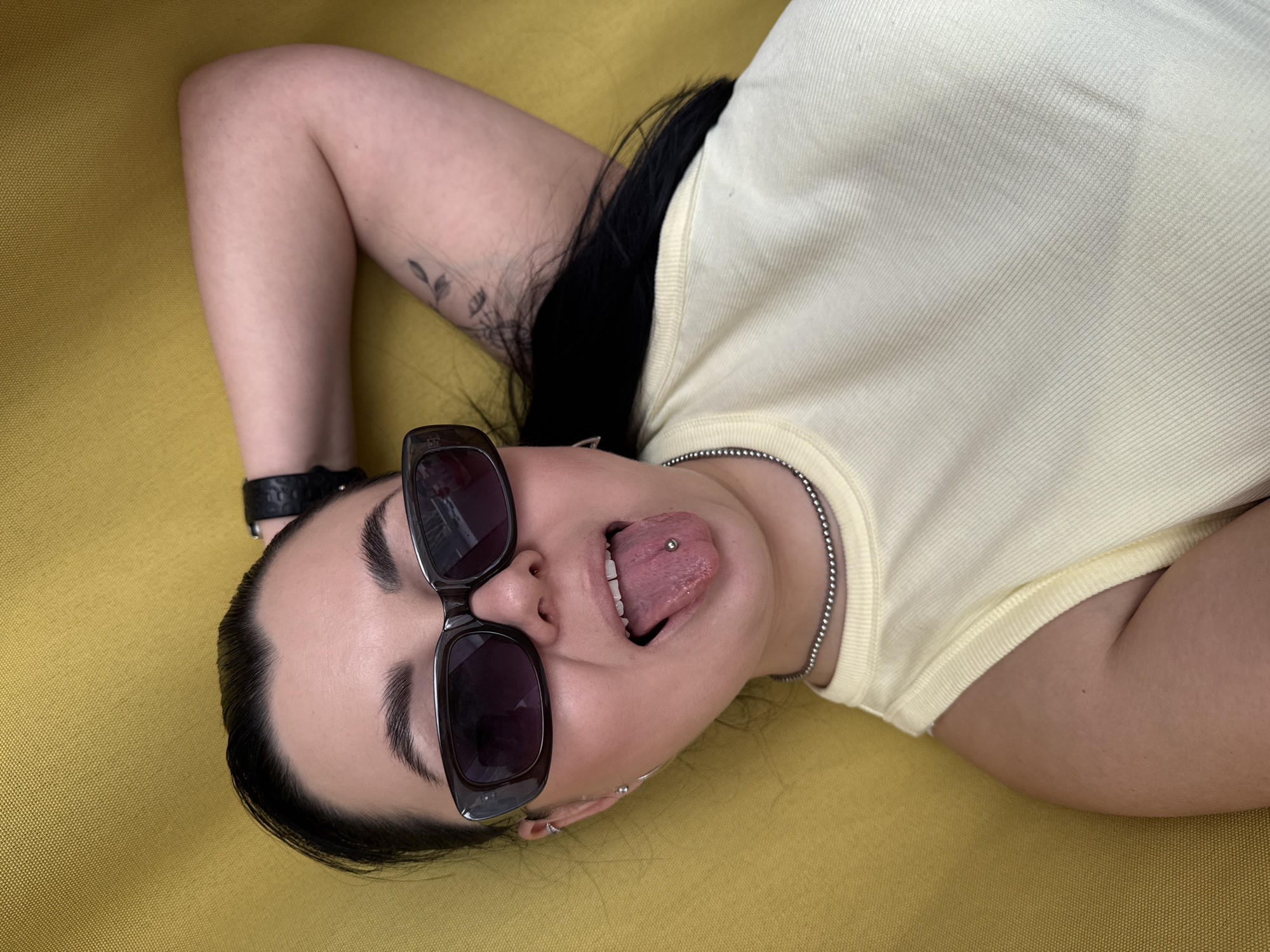 A person is lying on a yellow surface, possibly fabric. The person has dark hair pulled back and is wearing sunglasses. They are wearing a light yellow ribbed tank top and a silver ball-chain necklace. The person is sticking out their tongue, which has a piercing. A black watch is visible on the left wrist. A small floral tattoo is visible on the left upper arm. The setting is not discernible from the image, and a location in Chaikovsky, Russia, cannot be confirmed.