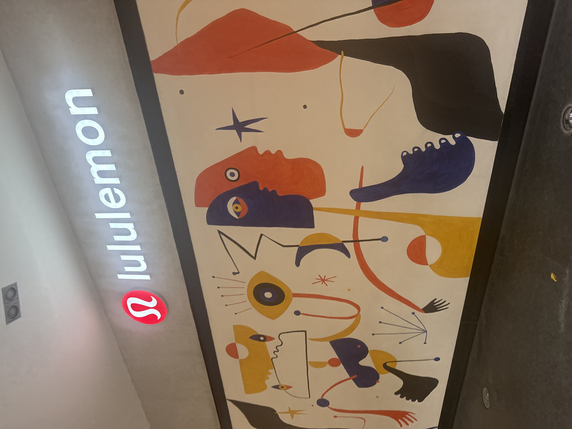 A vertically oriented photograph shows a Lululemon store with a large abstract mural on the wall next to the store sign. The sign, which reads "lululemon," is illuminated in white with the Lululemon symbol lit in red. The mural is a busy composition in the style of Joan Miró. It consists of a white background populated with colorful abstract shapes and lines in red, blue, yellow, black, and orange. These shapes suggest human forms, eyes, stars, and other organic elements. The photograph is taken from a slightly angled perspective, with the store's ceiling and the adjacent wall visible. Based on the Lululemon branding, this image is likely taken in a retail location, presumably in Puebla, Mexico.