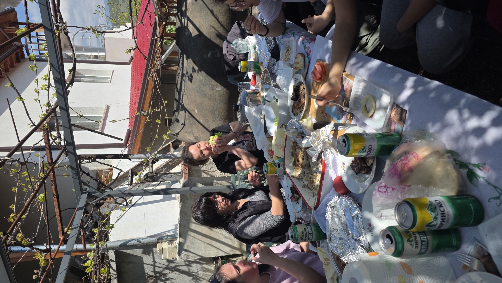 The image depicts a group of people enjoying a meal outdoors in what appears to be a backyard or patio setting. The setting is Katuntsi, Bulgaria. A white tablecloth covers a table laden with food, including meats, salads, bread, and cans of a local beverage. The people are seated around the table, engaged in eating and conversation. The background includes a building with a red roof and a trellis with budding vines. The lighting suggests it is daytime. The scene seems to capture a casual, communal meal in a private residence.