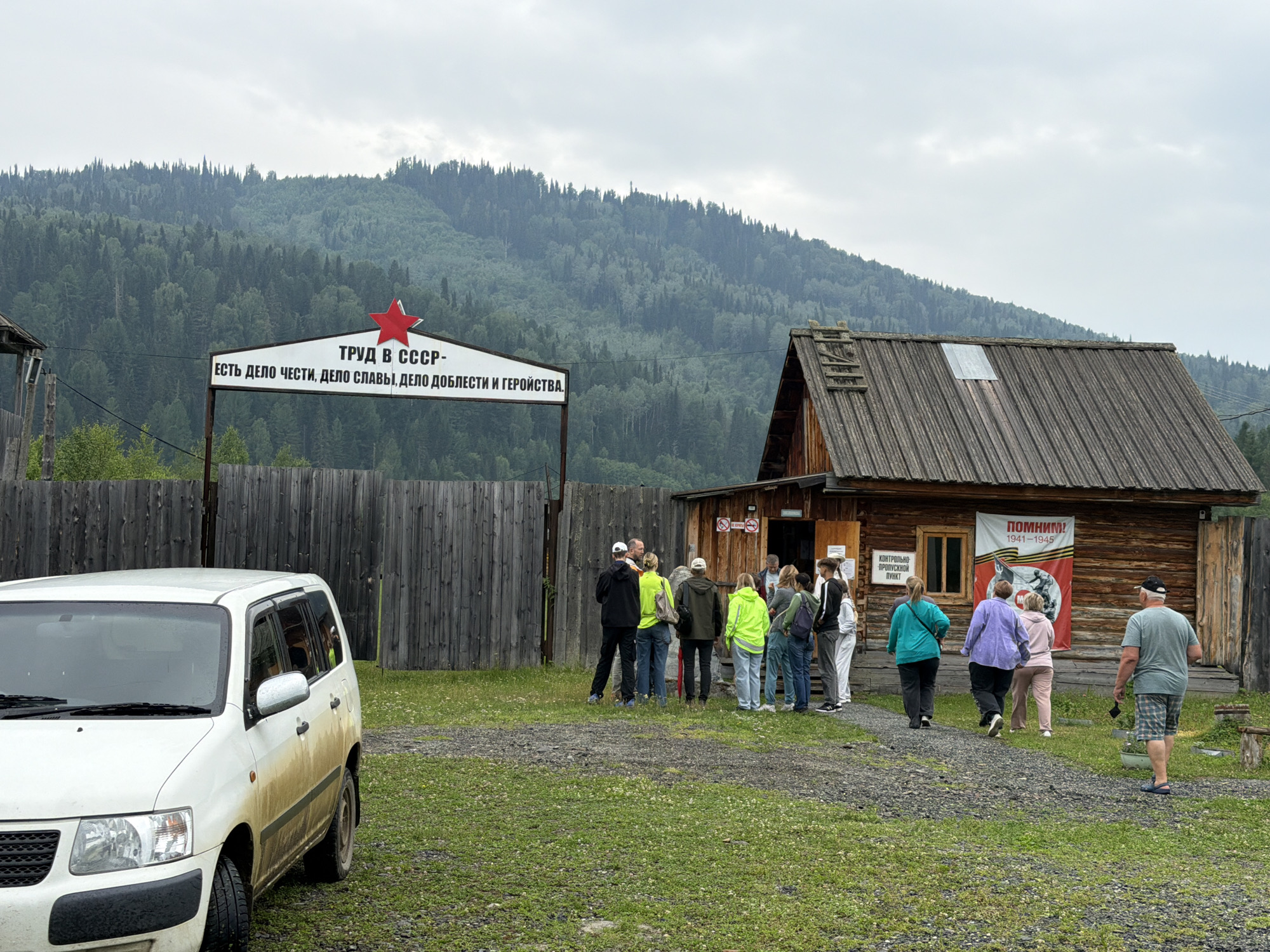 The image depicts an outdoor scene in Ust'-Kabyrza, Russia, featuring a wooden building with a rustic aesthetic and a group of people gathered outside. In the foreground, a white van, partially covered in mud, is parked on a gravel and grass area. A wooden fence runs along the left side of the scene, separating the area from a backdrop of a dense, forested mountain under an overcast sky.
A prominent sign with a red star is visible above the fence, displaying Cyrillic text that translates to "Labor in the USSR - is a matter of honor, a matter of glory, a matter of courage." To the right of the fence, the wooden building has a weathered appearance with a shingled roof. A banner hangs on the side of the building, featuring an image and Cyrillic text reading "We Remember! 1941-1945." Another sign with Cyrillic text is affixed to the wall near the entrance.
A group of approximately ten individuals, dressed in casual clothing, are positioned in front of and near the entrance of the building. Some are facing the building, while others are engaged in conversation. The overall atmosphere appears to be one of a gathering or a visit to a historical site. The environment is rural, characterized by the natural landscape and simple structures.