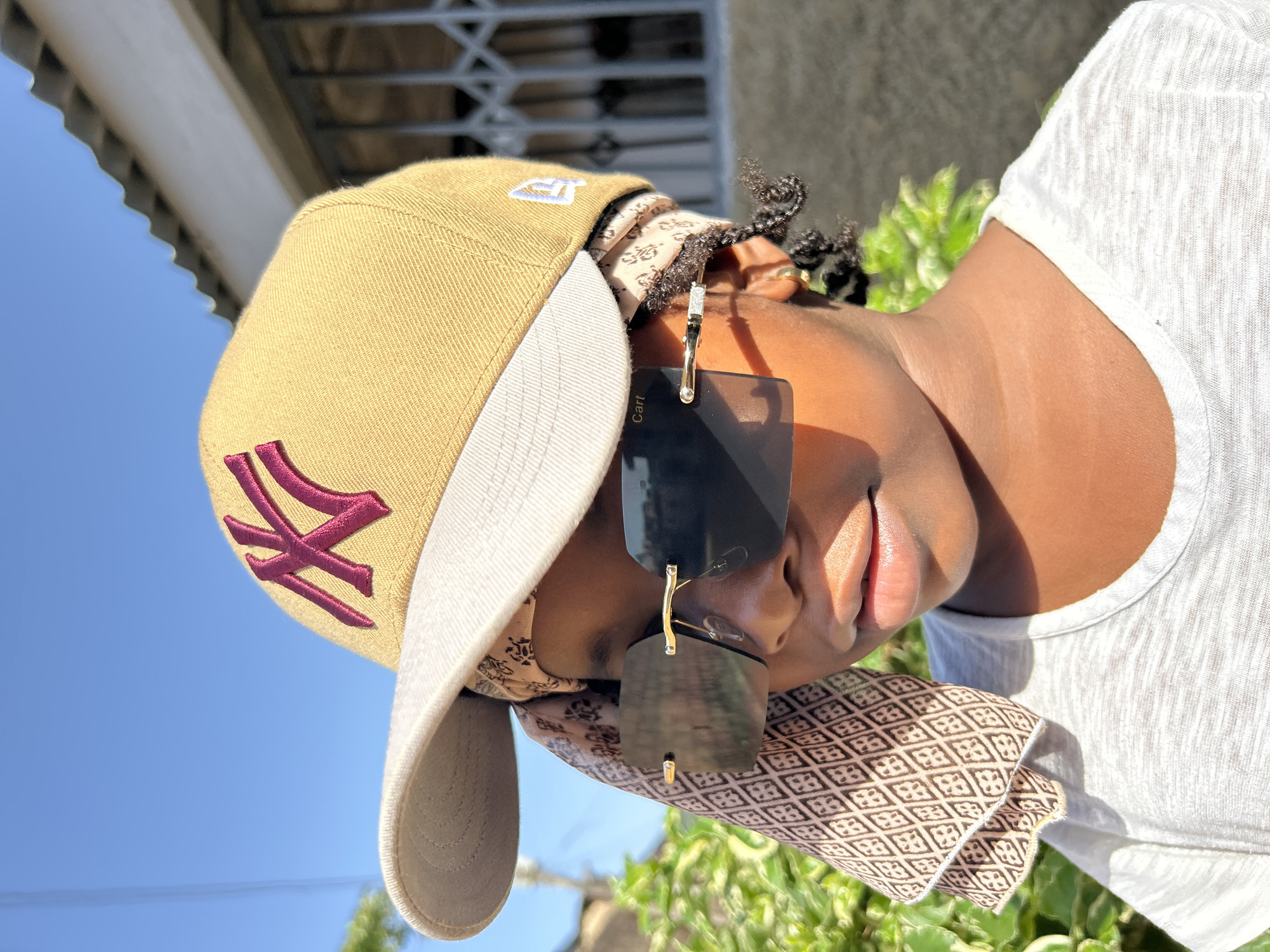 A person with dark skin wears sunglasses and a cap with a NY logo, outdoors in Jos, Nigeria. The person has braids and wears a patterned headscarf under the cap and around their neck, coupled with a white t-shirt. The background includes greenery and a building structure under a clear sky.