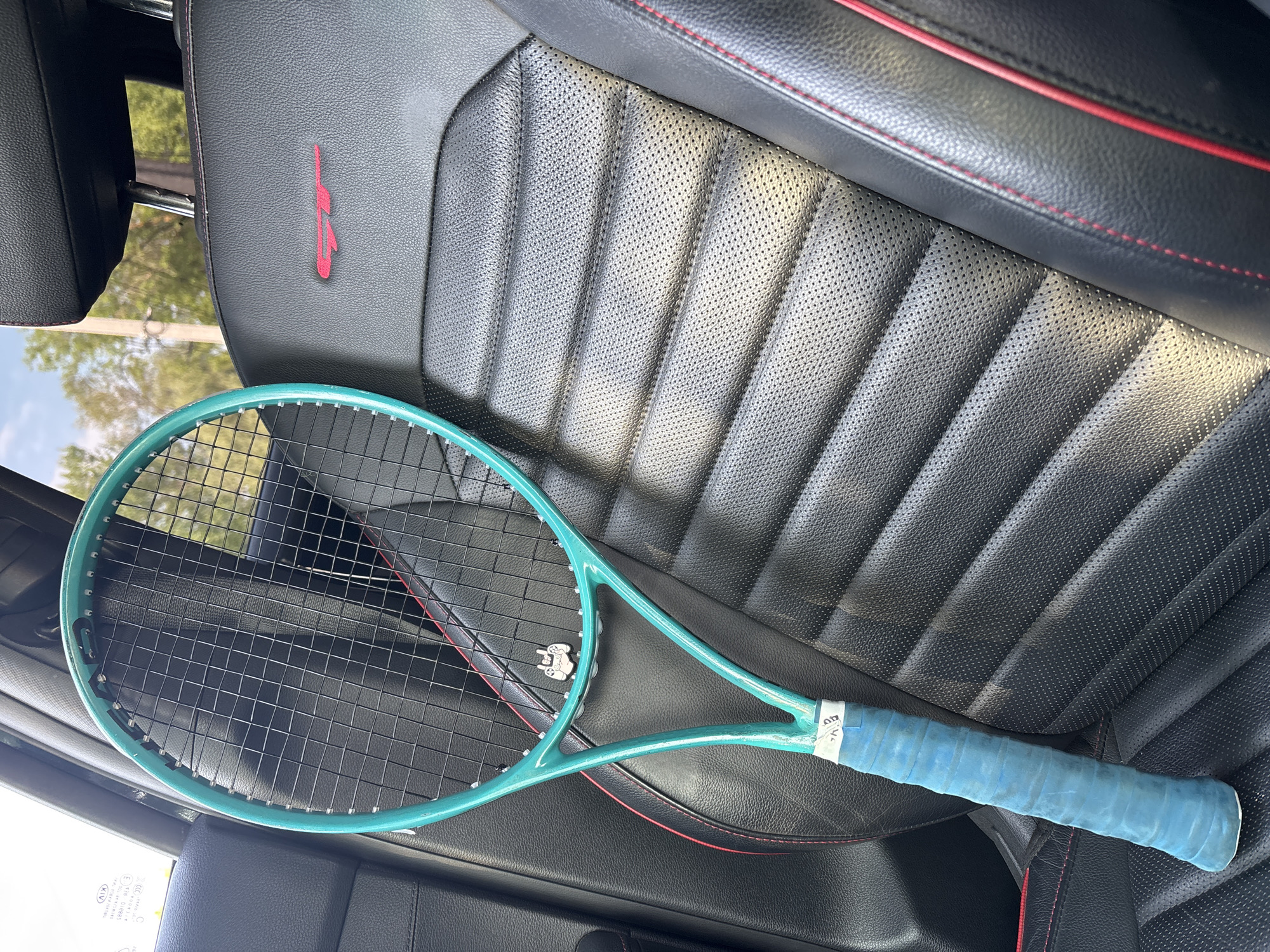 A light blue tennis racket lies on a black leather car seat with red stitching. The seat has a perforated pattern and a red logo, which appears to be the letters "GT". The handle of the racket is wrapped in a worn light blue grip. The background shows a glimpse of trees and sky through the car window. The image seems to be taken inside a car, possibly in Chaikovsky, Russia.