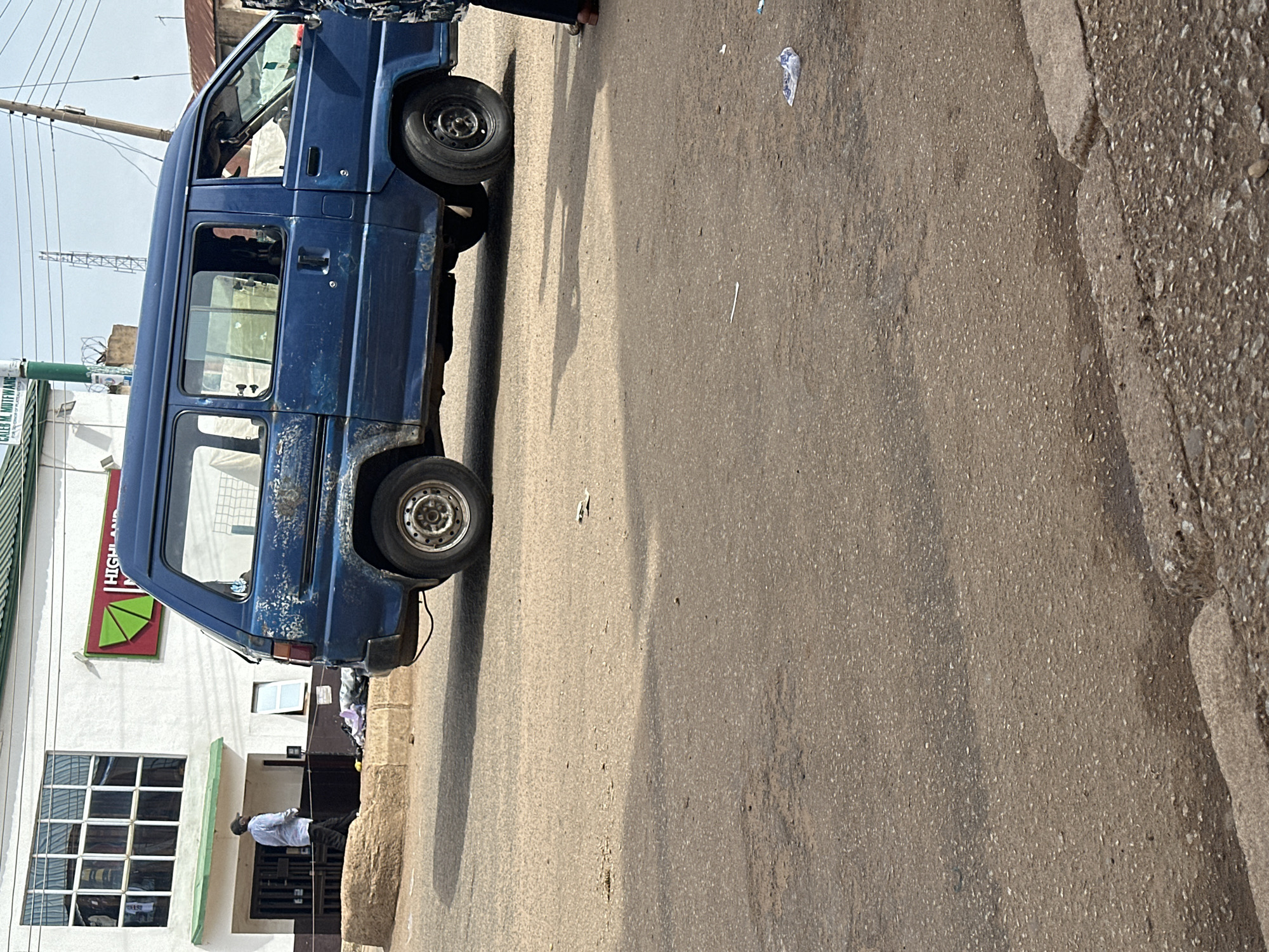 In Bukuru, Nigeria, a blue van is parked on the side of a paved road. The van is old and shows signs of wear, with rust and chipped paint. The road surface is rough and cracked in places, with some debris scattered on it, including a plastic bag. To the left of the van, a building with a white facade and a green awning is visible. The building has large, multi-paned windows. A man in a light-colored shirt and dark pants is walking away from the building towards the van. He is wearing a cap. The environment appears to be an urban street scene, with a clear sky overhead.