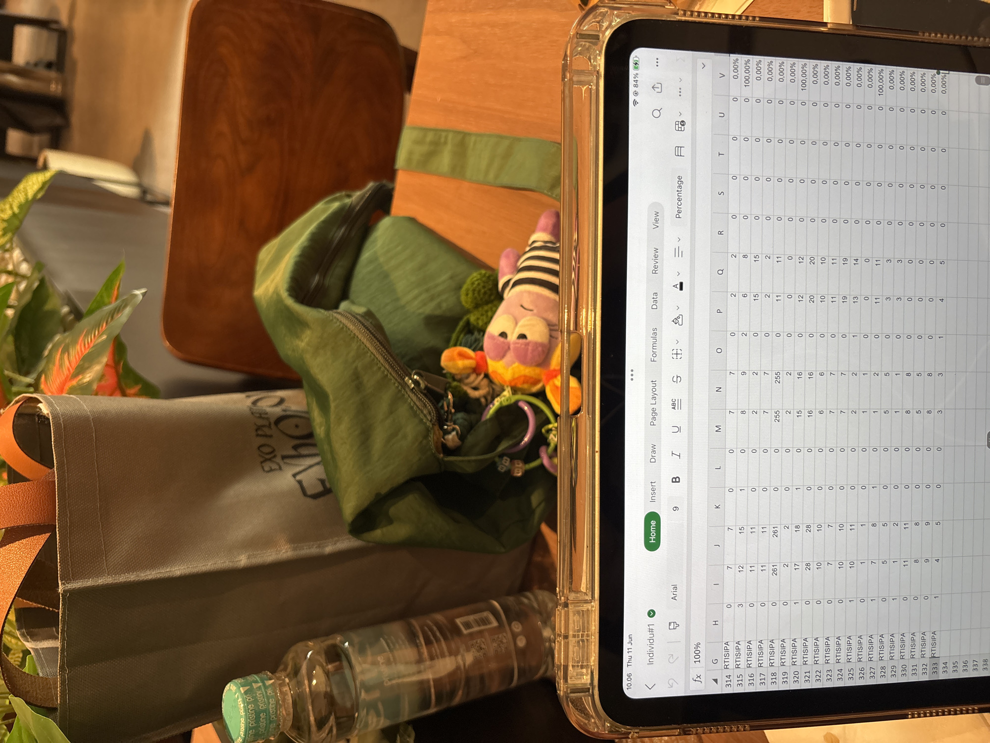 The image shows a close-up view of a tablet screen displaying a spreadsheet, likely in a coffee shop or office setting. The spreadsheet contains columns labeled "G" through "V" with numerical data and percentages. The surrounding environment features a green tote bag with orange straps, a water bottle with a green cap, and a stuffed toy. The tablet is placed on a wooden surface, possibly a table. The time stamp on the tablet indicates "10.06 Thu 11 Jun." The scene suggests a person is working or studying on their tablet while surrounded by personal belongings. There is no visible indication of the location being in West Jakarta, Indonesia.