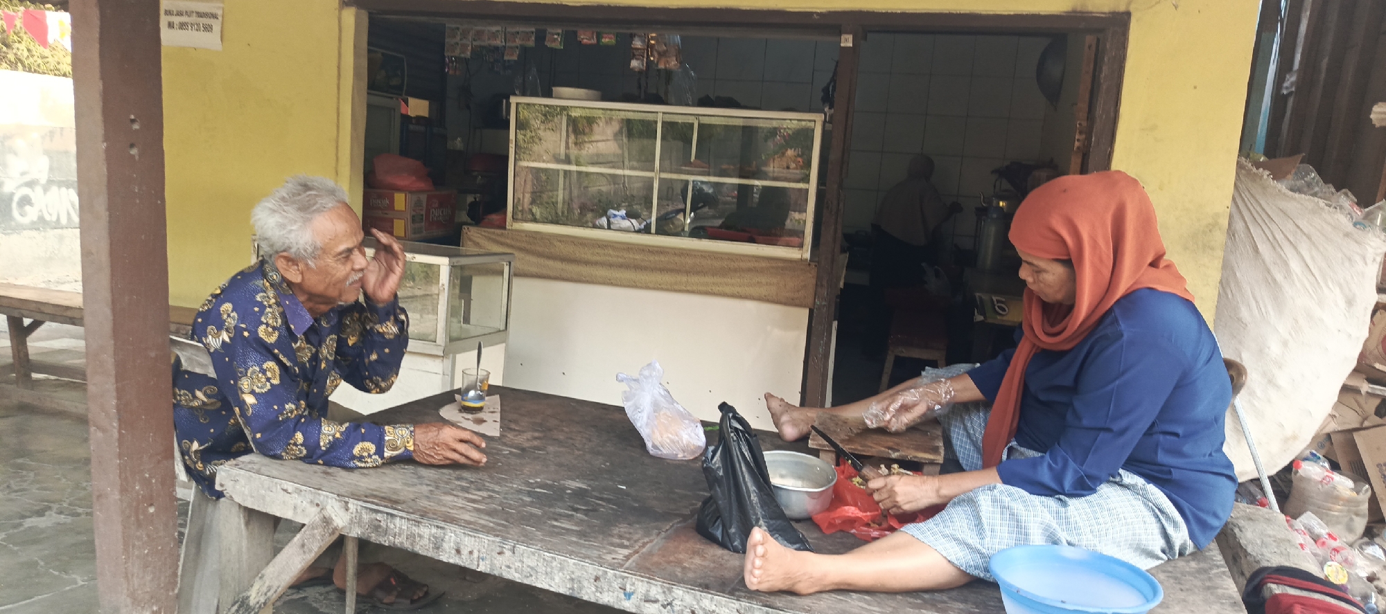 In Sukaresmi, Indonesia, two individuals are seated at a weathered wooden table in front of a small shop. An elderly man with grey hair, wearing a blue patterned shirt, sits on the left, holding his hand to his face. On the right, a woman wearing an orange headscarf and a blue long-sleeved shirt is seated, preparing food with a knife and plastic-wrapped ingredients. A variety of items, including plastic bags, bowls, and what appear to be bottles, are scattered on the table and the surrounding ground. The shop behind them has a glass display case filled with items, and shelves stocked with products. The overall environment suggests a casual, outdoor setting associated with a local business.