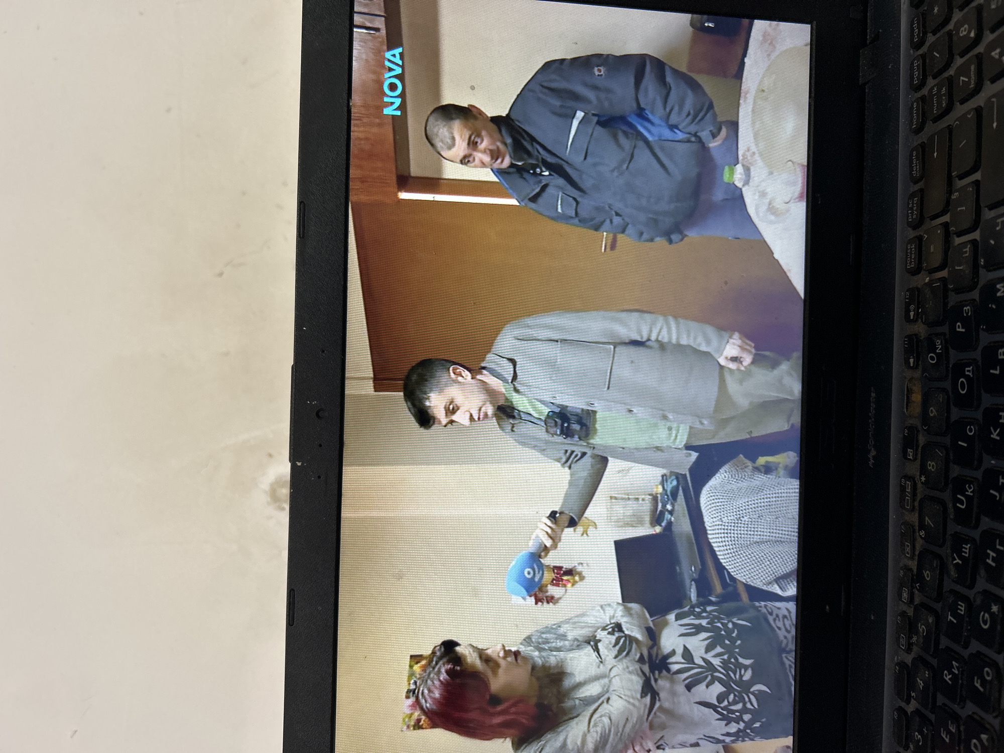 The image captures a black laptop screen displaying a television broadcast, with the laptop's Cyrillic and Latin character keyboard partially visible in the foreground. The broadcast content originates from the Bulgarian channel "NOVA," identifiable by its logo on the upper right of the screen and on a microphone. The scene is an interior room, presumed to be a residential setting in Sofia, Bulgaria, characterized by light-colored walls and a visible wooden doorframe to the right.
Three individuals are present on the screen. On the left, a woman with reddish-brown hair wears a patterned light-colored top, standing with crossed arms and looking towards the center. In the middle, a man with dark, short hair, dressed in a light green shirt and a light gray jacket, holds a blue "NOVA" branded microphone towards the woman. A recording device is suspended around his neck. On the right, a second man with very short hair, wearing a dark blue and gray puffer jacket, stands with hands in his pockets, observing the interaction.
Behind the woman, a colorful framed picture hangs on the wall. To the left of the central man, a dark laptop sits on a surface, partially obscured by a white patterned fabric. A light-colored, circular table is visible in the lower right portion of the screen, bearing a green-capped plastic bottle and a small glass with a reddish liquid.