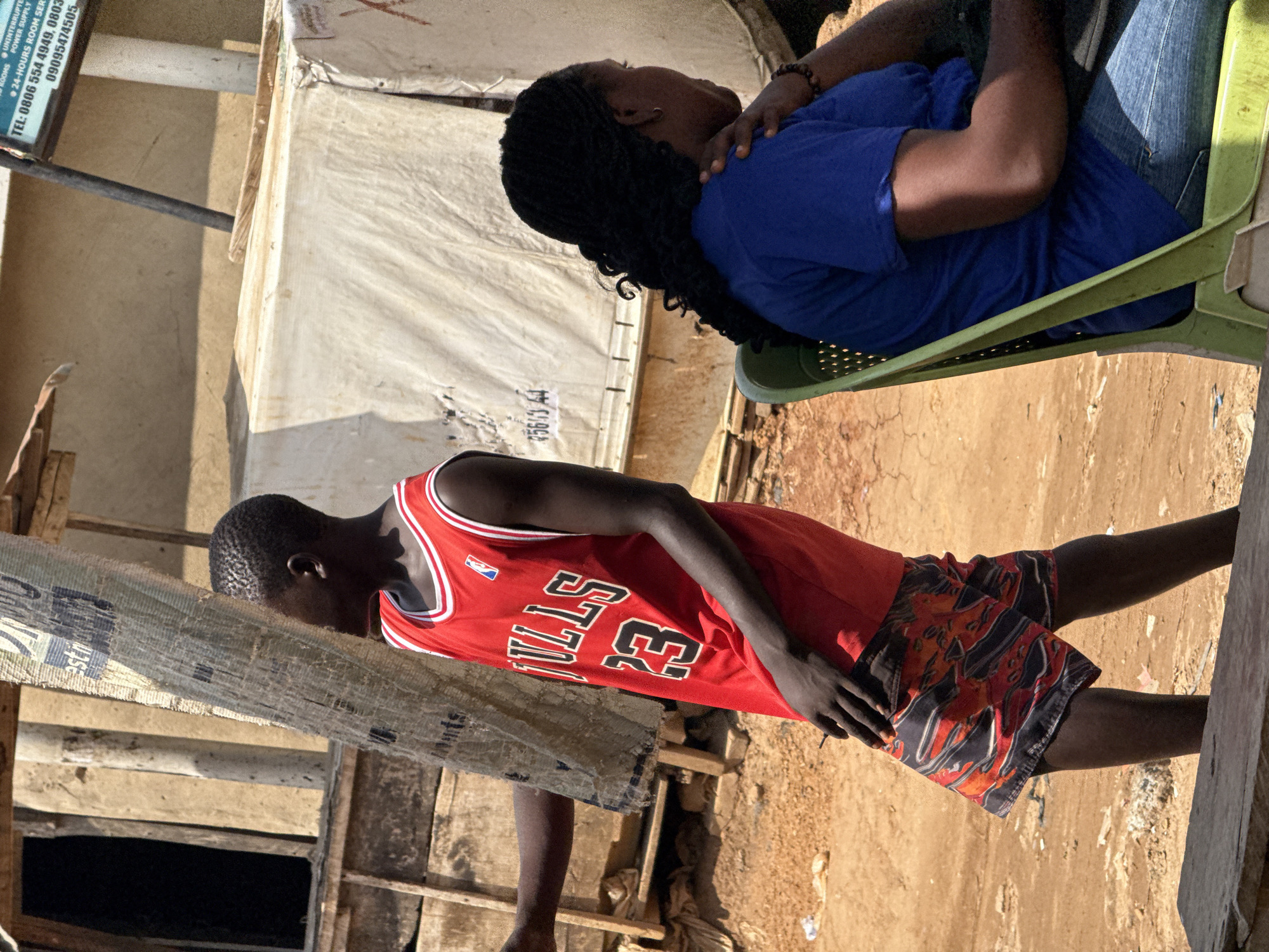 In a dusty, outdoor setting in Abuja, Nigeria, a young Black male wearing a red Chicago Bulls basketball jersey with the number 23 and patterned shorts stands to the left. He is partially obscured by a large, textured vertical pole made of woven material. To his right, a Black female with braided hair is seated on a green plastic chair, wearing a blue t-shirt and denim jeans. She is looking to the right, with her right hand resting on her shoulder. Behind them, there are various structures, including wooden scaffolding, a white tarp, and some debris scattered on the ground. The environment appears to be a street or market area, with dry, reddish-brown earth underfoot.