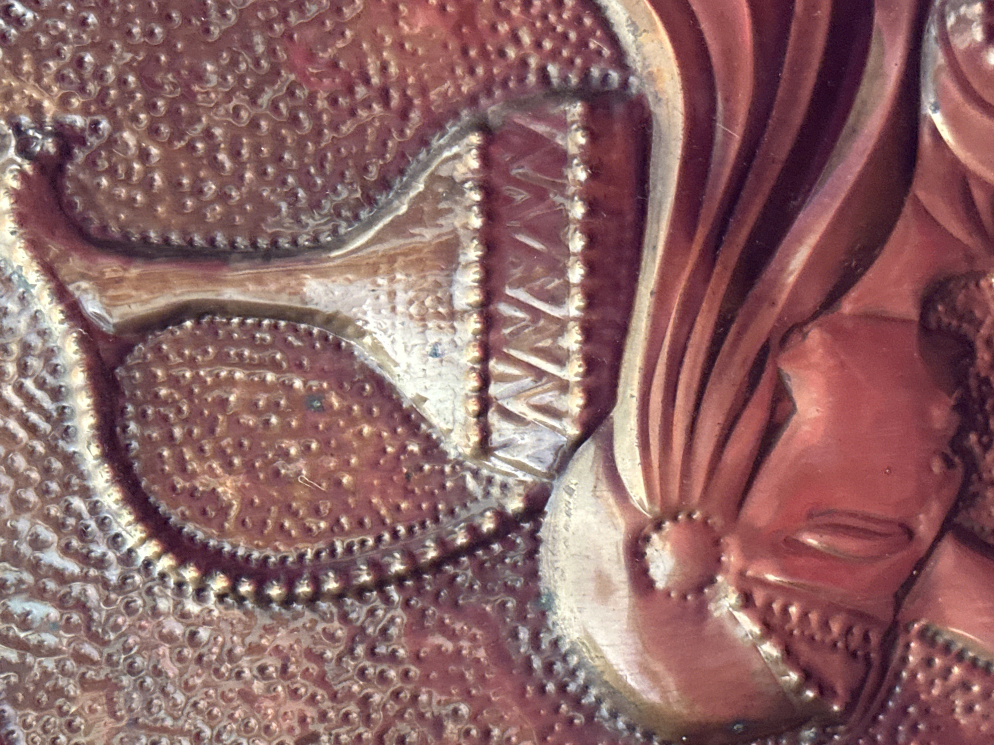This is a close-up, cropped image of a relief sculpture with a metallic, coppery sheen. The sculpture depicts a stylized human profile, likely female, with closed eyes and flowing hair. Above the profile, a decorative element resembling a fluted vase or an urn with a patterned base is visible. The background is textured with numerous small, raised circular indentations. The overall aesthetic suggests an Art Nouveau or similar decorative style. The material appears to be metal, possibly copper or brass, with some areas showing oxidation or patina. The image is focused on the texture and form of the sculpture itself, with no environmental context provided that would indicate a specific location in Nevinnomyssk, Russia, or any other city.