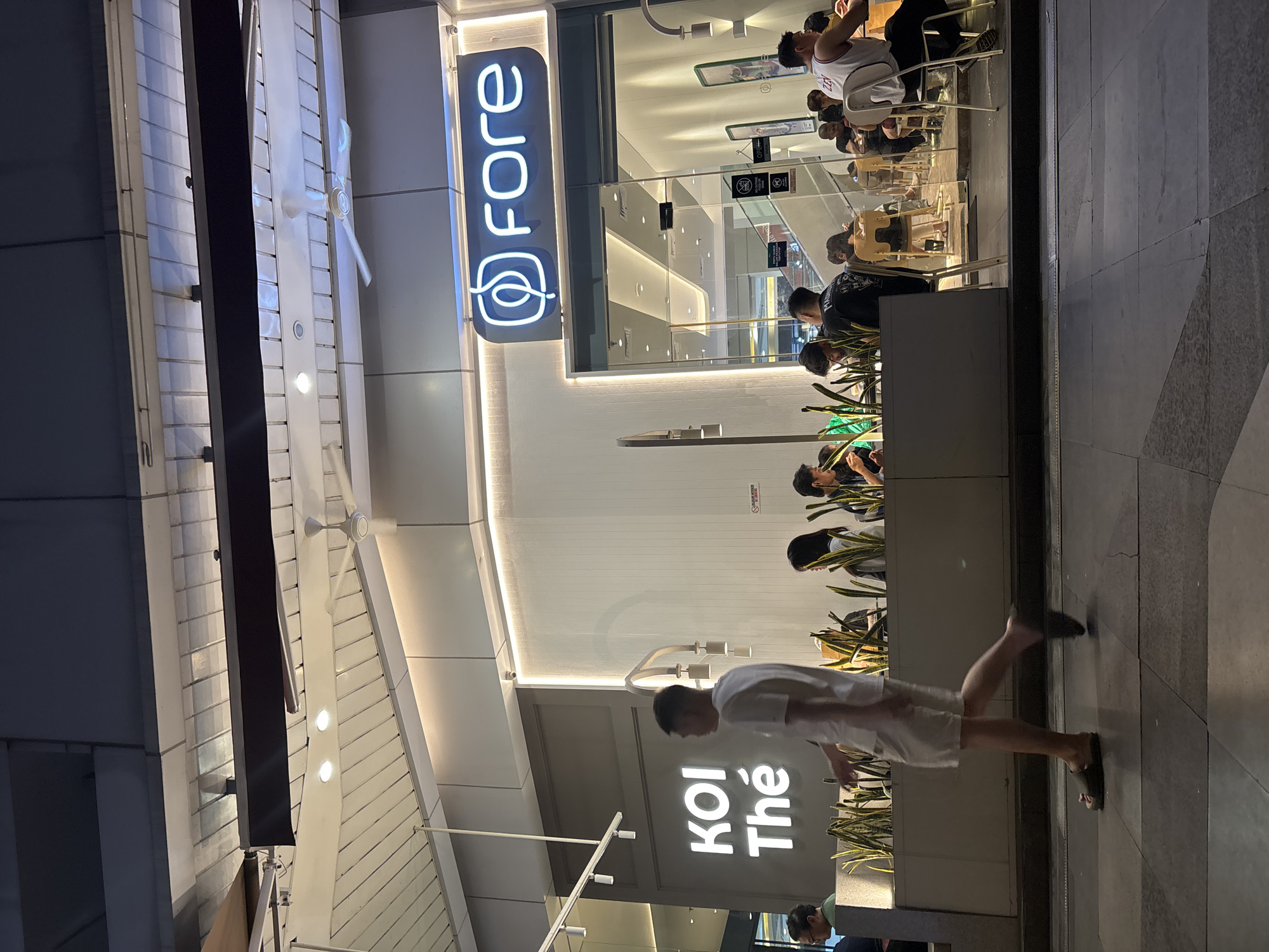 The image depicts the exterior of a cafe, identified by signage as "KOI Thé" and "fore," located in North Jakarta, Indonesia. The cafe appears to be situated in a commercial or mall area, with large glass windows offering a view of the interior where customers are seated at tables. A ceiling fan is visible, suggesting an indoor or covered outdoor seating area. A man in white shorts and a white t-shirt is walking past the cafe on a paved walkway, in the foreground and to the left of the frame. The lighting suggests it is evening or nighttime. The overall scene is that of a typical urban commercial establishment.