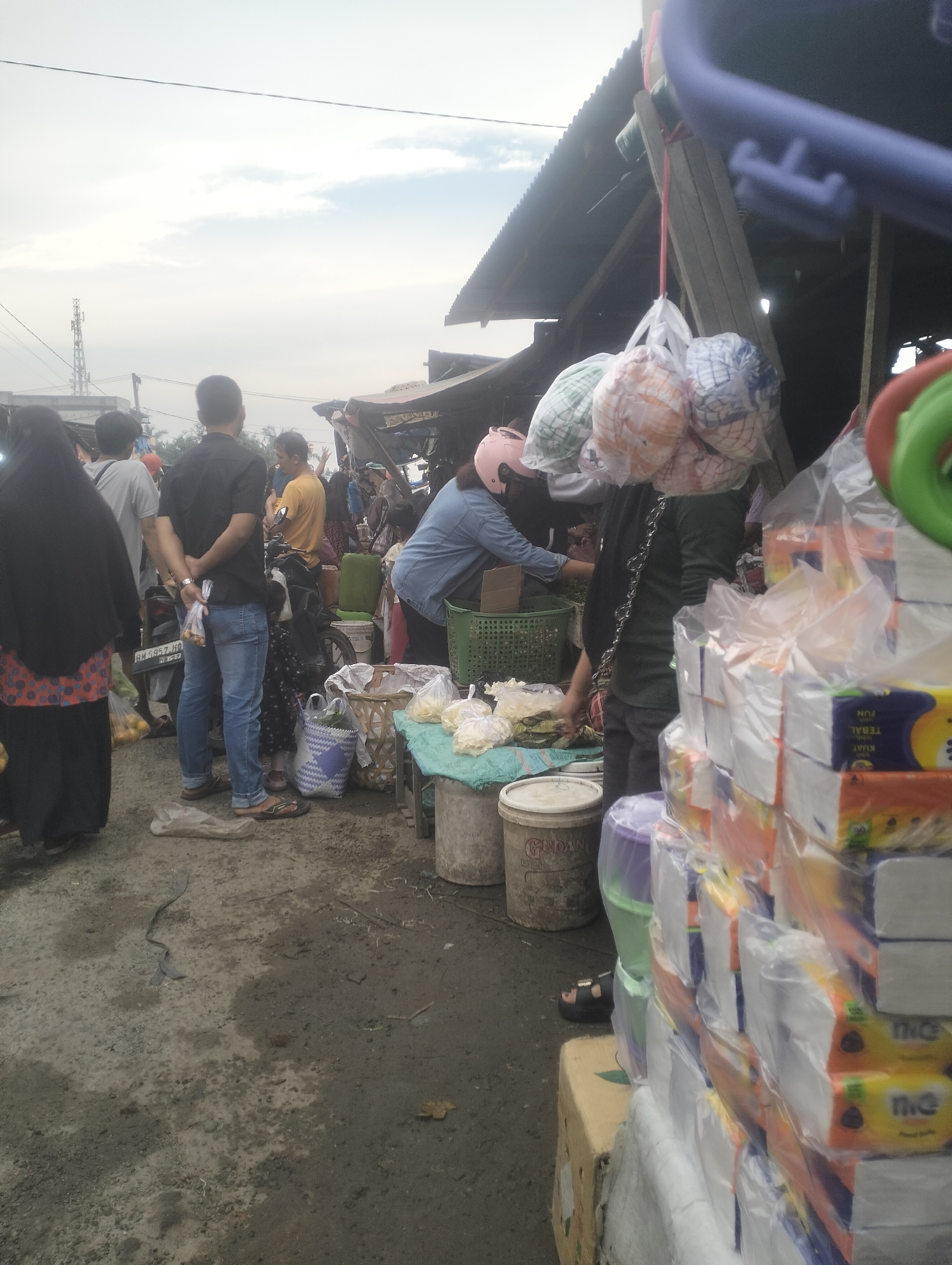 The image depicts a bustling outdoor market scene, likely in Dumai City, Indonesia. A variety of people are present, some dressed in traditional Islamic attire and others in casual clothing. In the foreground, a person wearing a black hijab and a patterned skirt is standing. To their left, a man in a black shirt and blue jeans is seen from the back, holding a plastic bag. A motorcycle is parked behind him.
Several stalls are set up, with goods displayed on tables and in baskets. Items such as packaged tissues, what appear to be snacks or food items, and colorful textiles are visible. Some of the goods are wrapped in plastic for protection. In the background, a tall communication tower can be seen, suggesting an urban or semi-urban environment. The market appears to be on a dirt or gravel surface, with some puddles indicating recent rain or a damp environment. The sky above is overcast. The overall impression is one of a lively and active marketplace.